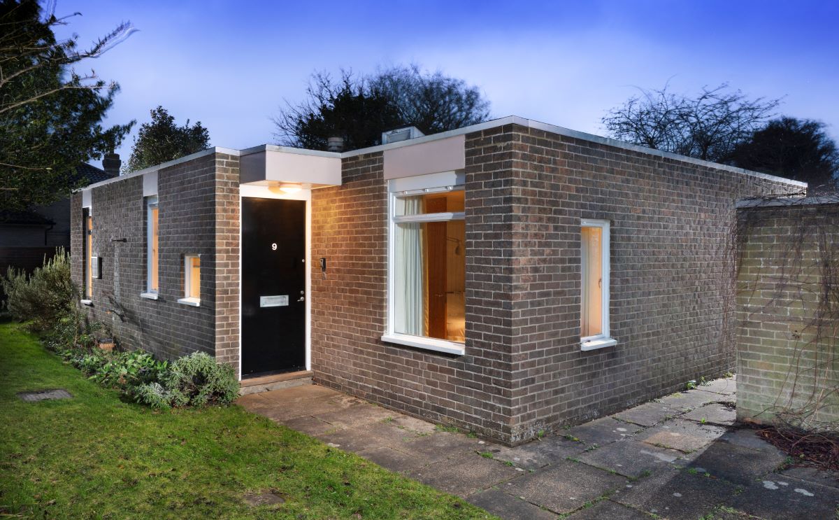 A single storey brick bungalow with lighted windows and a black door.