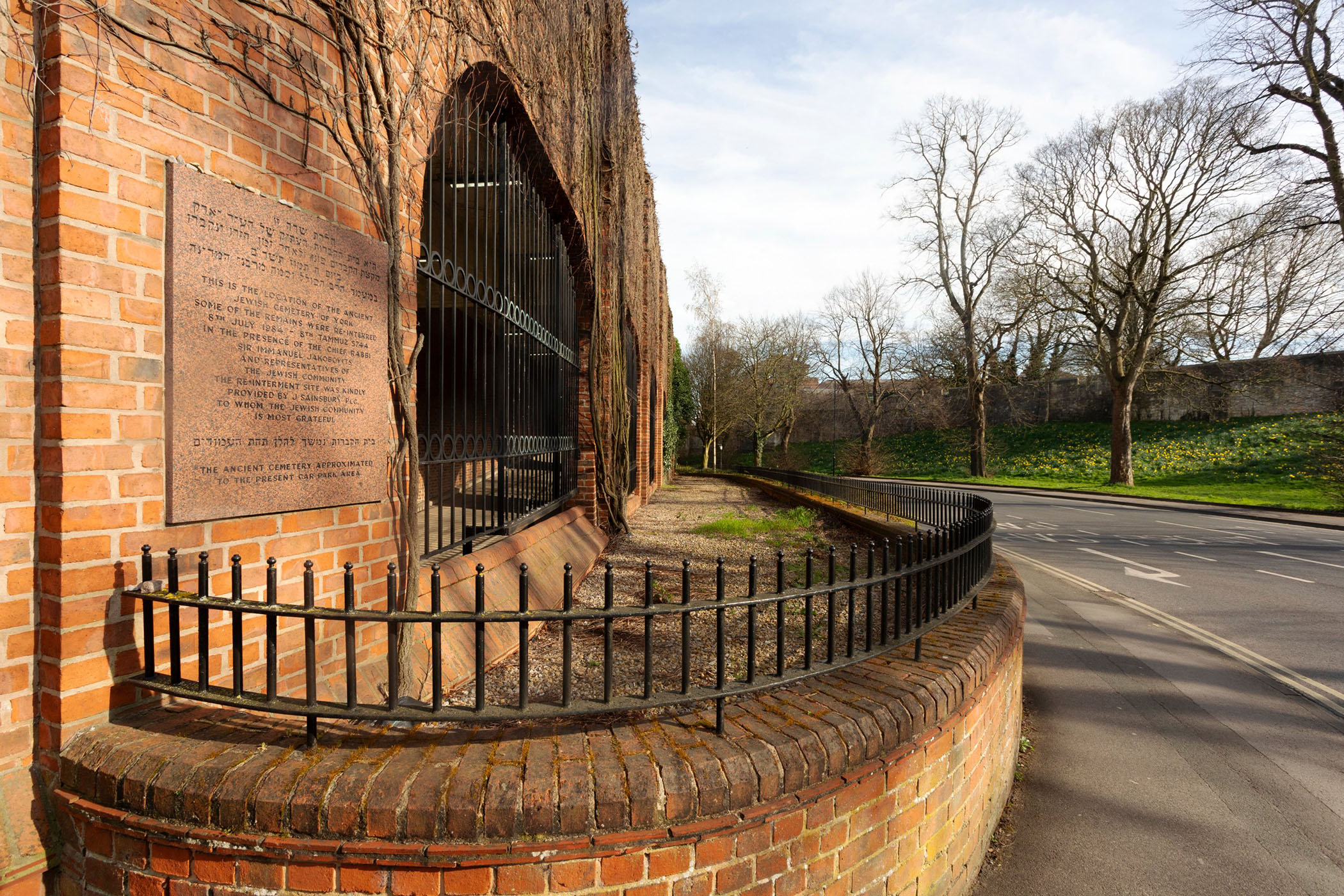 A view of a stone memorial plaque on the side of a brick building, with a fenced-off gravel area and a road alongside