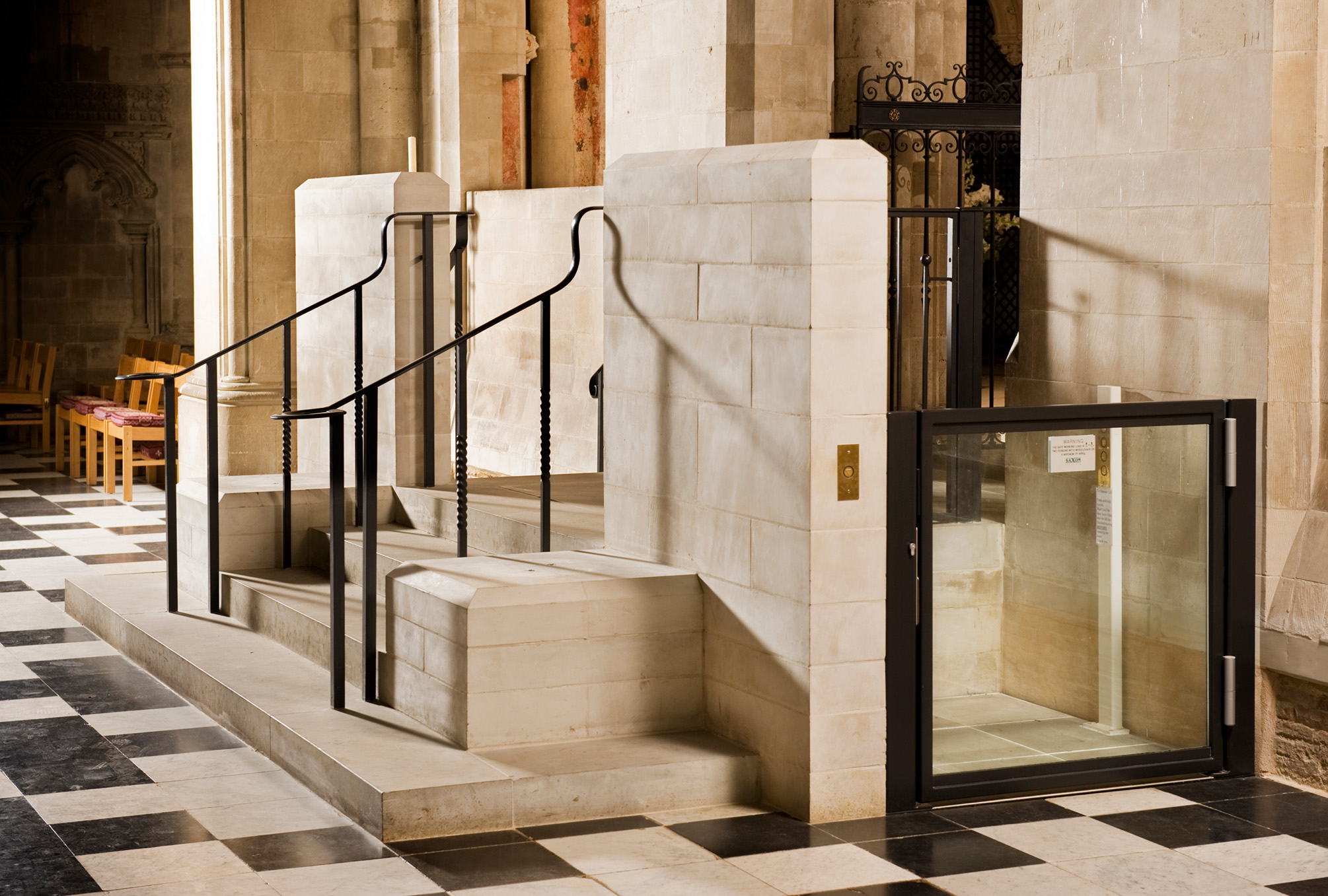 Interior view showing a wheelchair lift built within a cream stone staircase with black railings. The ground floor is black and white tiles.