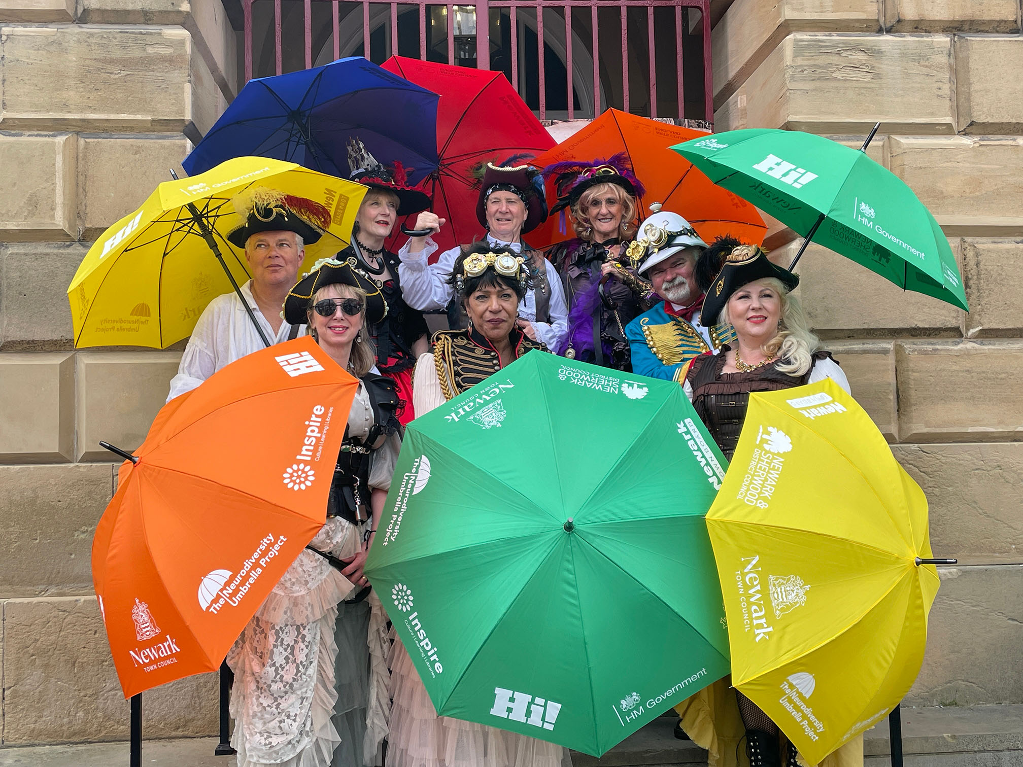 A group of people wearing costumes and holding colourful umbrellas stood on steps flanked by stone walls
