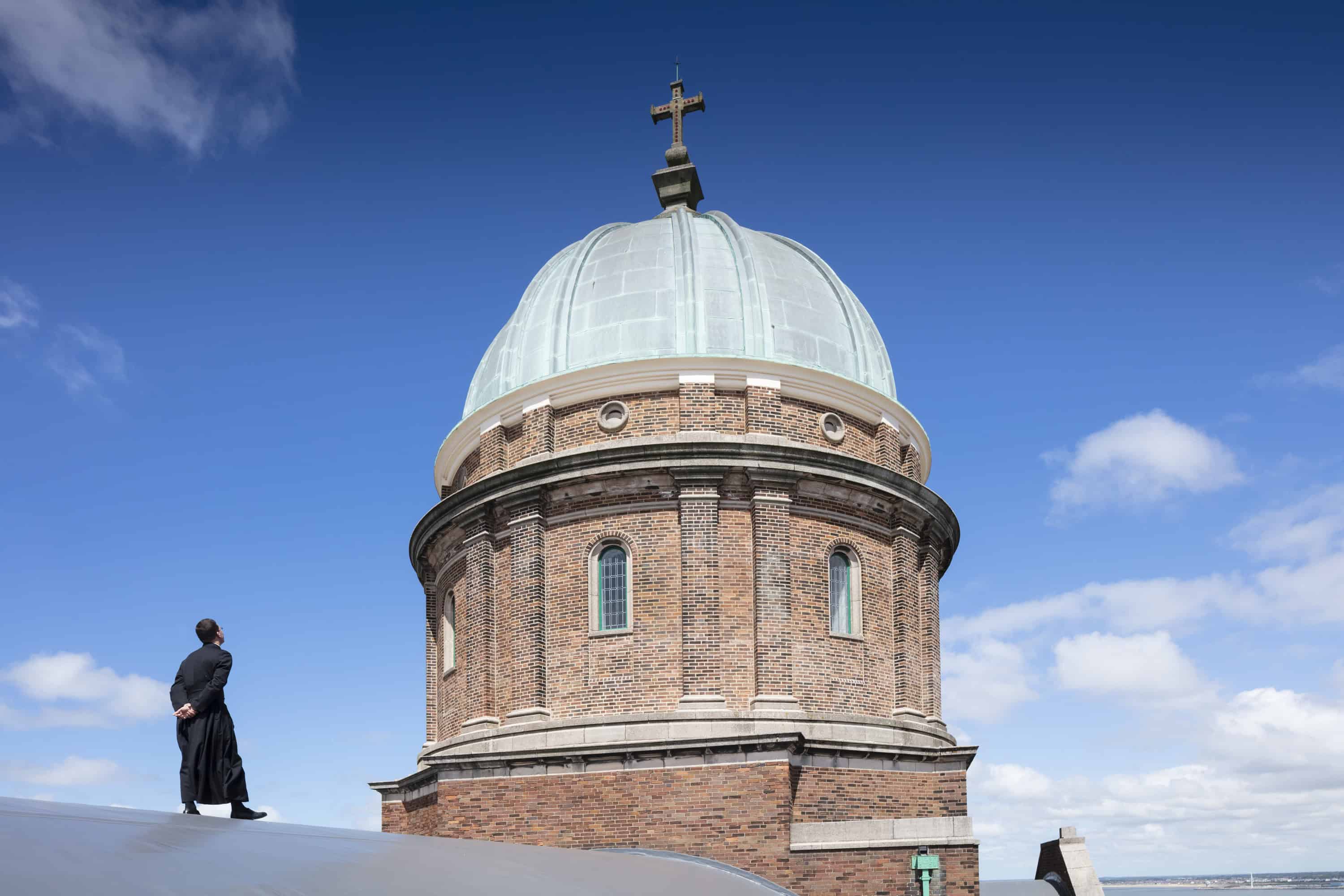 Image of the top of a church, featuring a dome with a cross at the top. There is a man in black robes in front of the dome. 