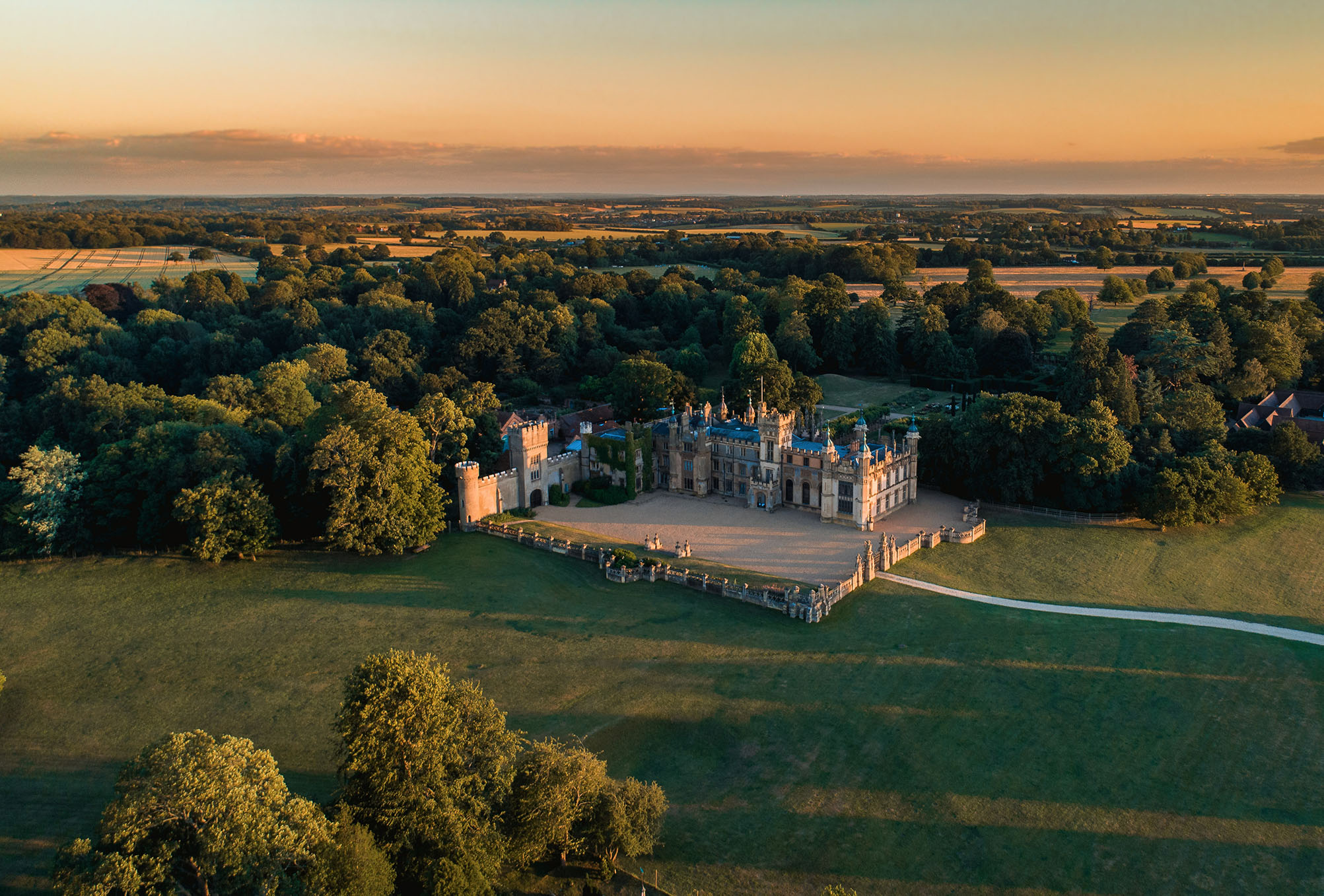 An aerial photograph shows the Knebworth House and Garden surrounded by trees with fields in the distance and a setting sunset.