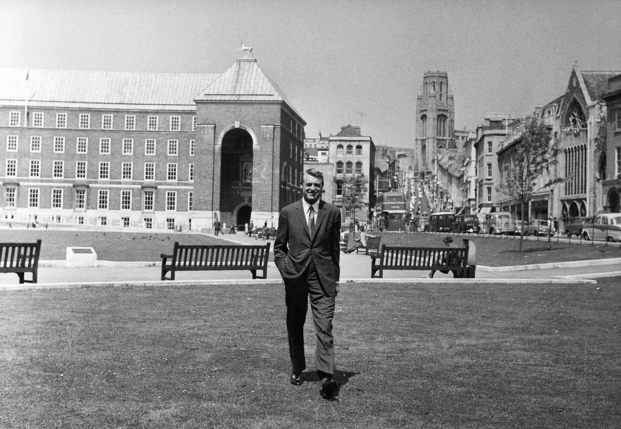 A black and white photo of the actor Cary Grant stood in the middle of a grassy area surrounded by park benches, cars and buildings