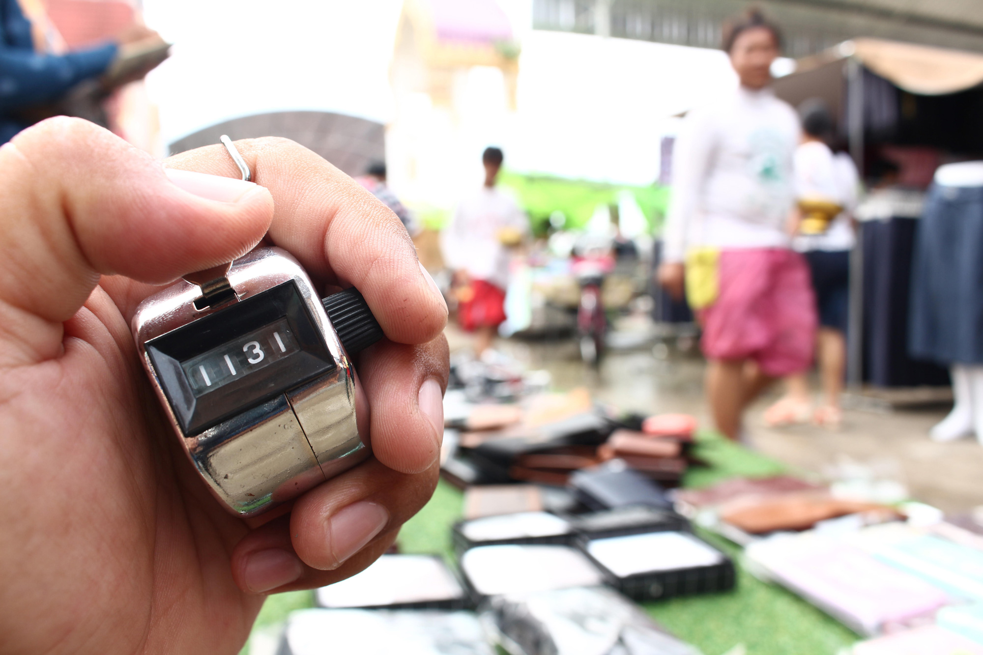 Hand holds a clicker counting device on left of the image. In the blurred background on the right are people and products at a market. 