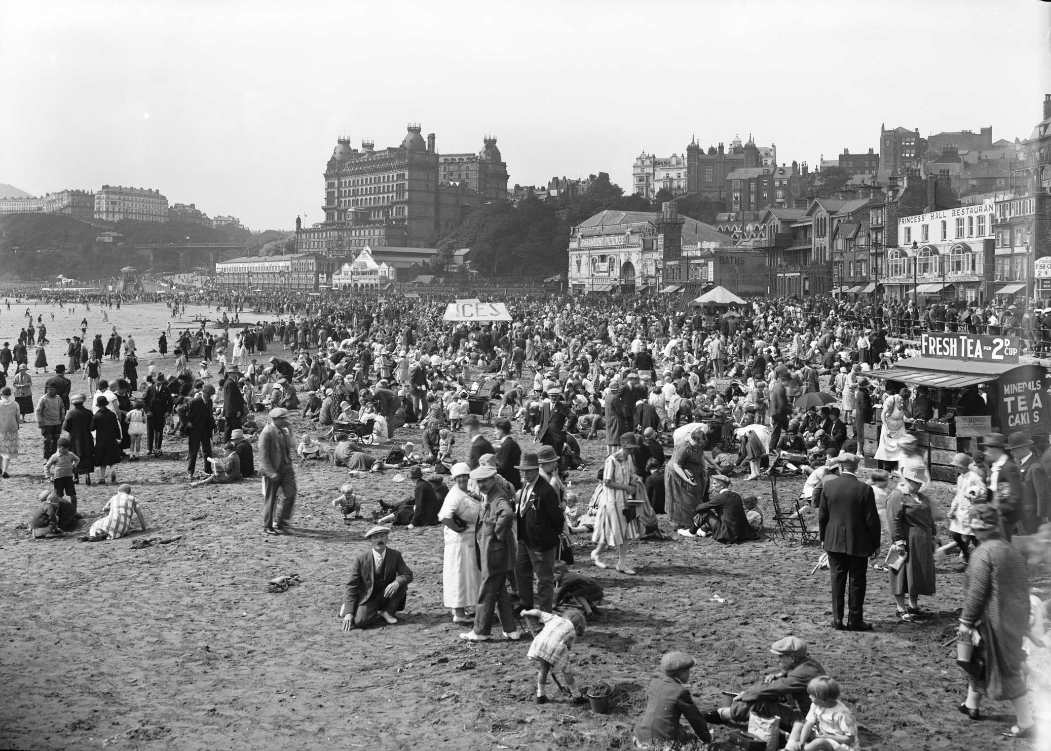 An archive photograph of a seaside resort beach scene with crowds in 1920s clothing.
