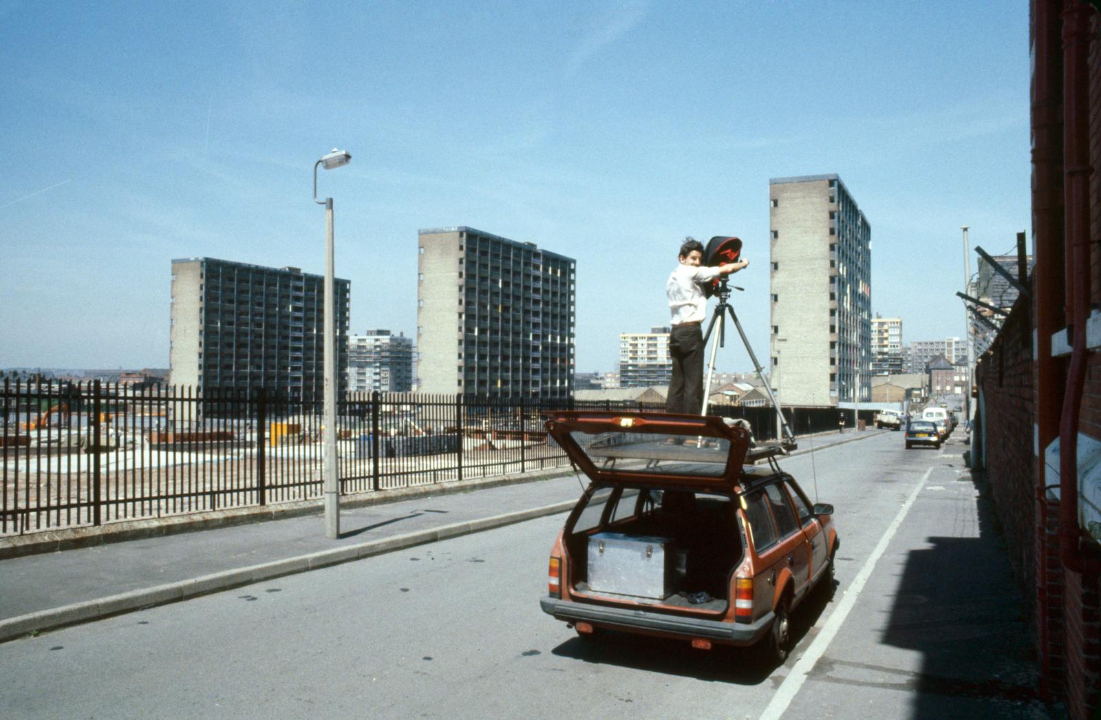 Photographer and tripod set up on the roof of a parked car on a quiet street. The camera points towards three tower blocks in the distance.