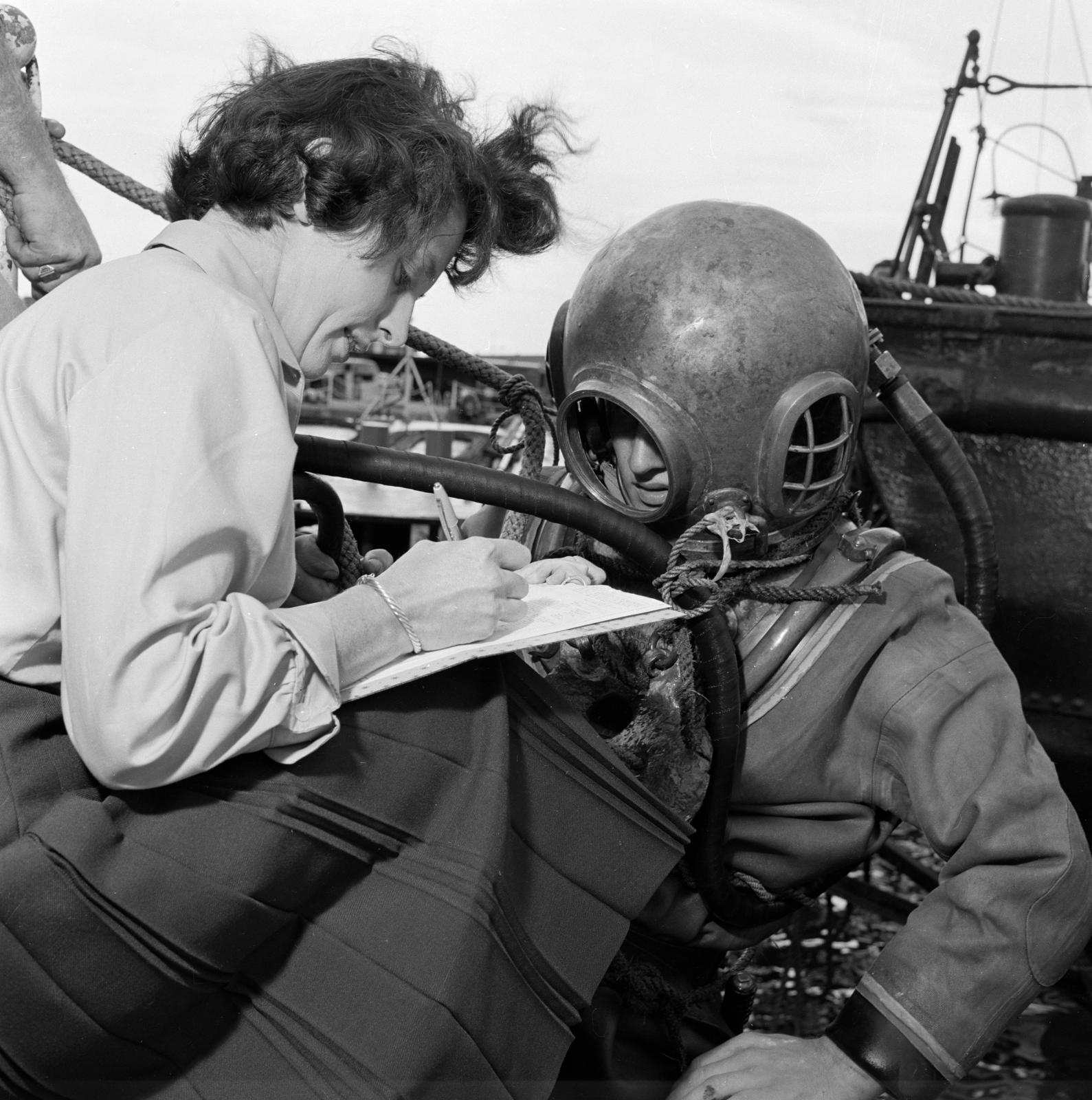 A black and white photo of a female gallup pollster with a clipboard surveying a man wearing a deep sea diving suit.