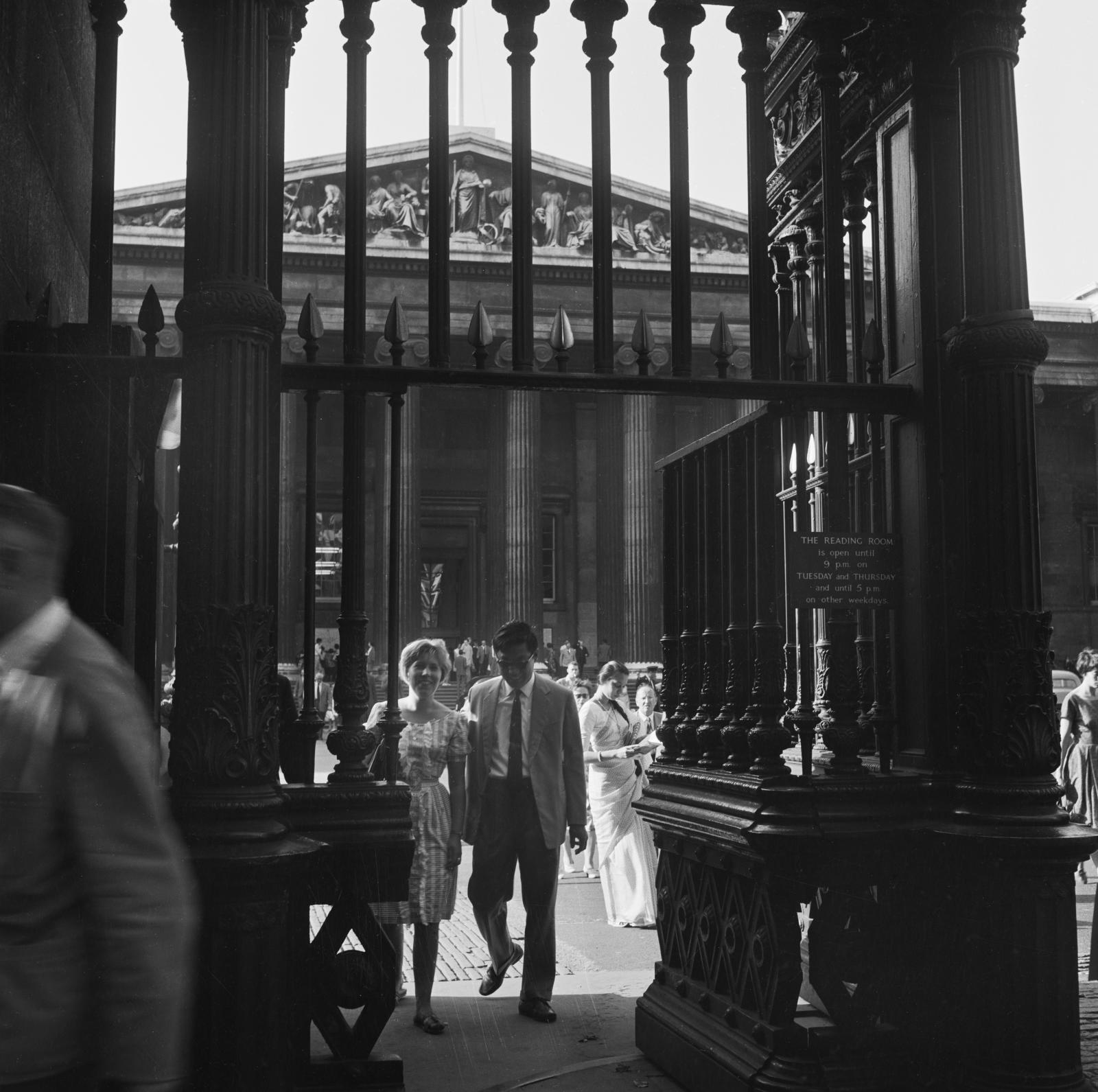 A couple make their way through wrought iron gates leaving the British Museum's south entrance while behind them crowds are milling about in the museum's forecourt.