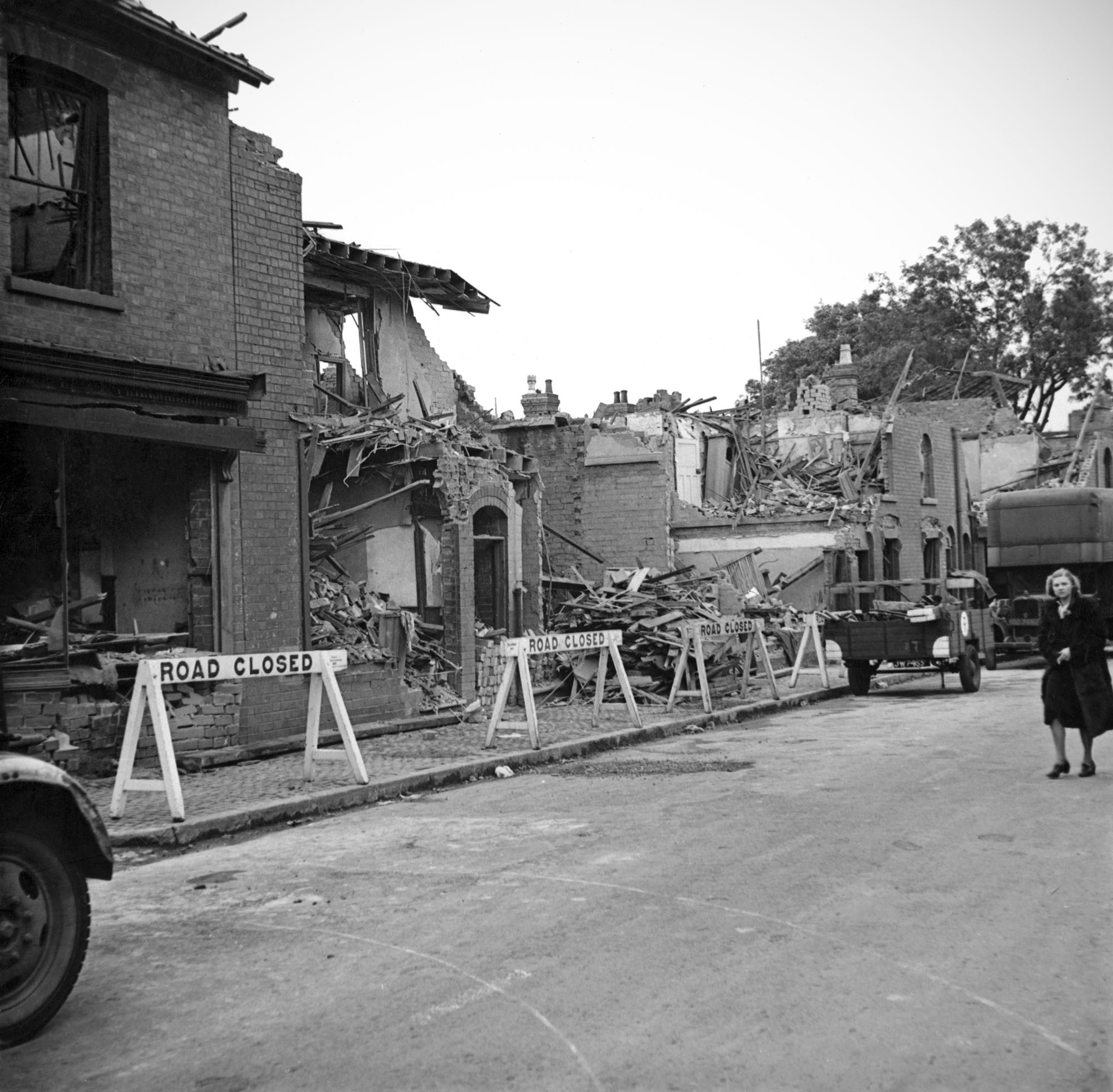 In front of the damaged houses are 'road closed' signs and a woman walking along in the road.