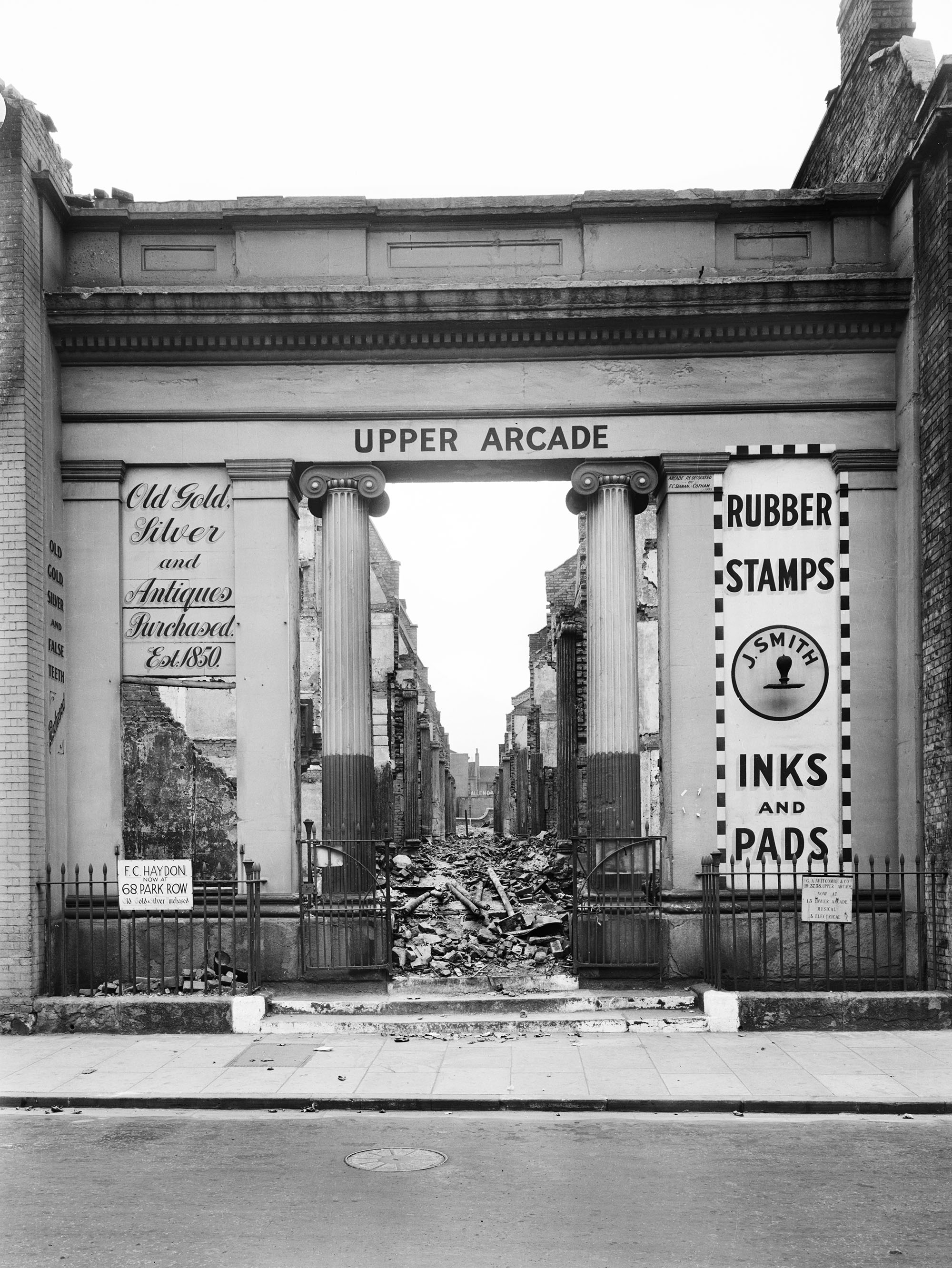 Shop signs at the entrance to the arcade read: 
On the left hand side: "Old Gold Silver and Antiques Purchased. Established 1850."
On the right hand side: "RUBBER STAMPS INKS AND PADS. J. SMITH"