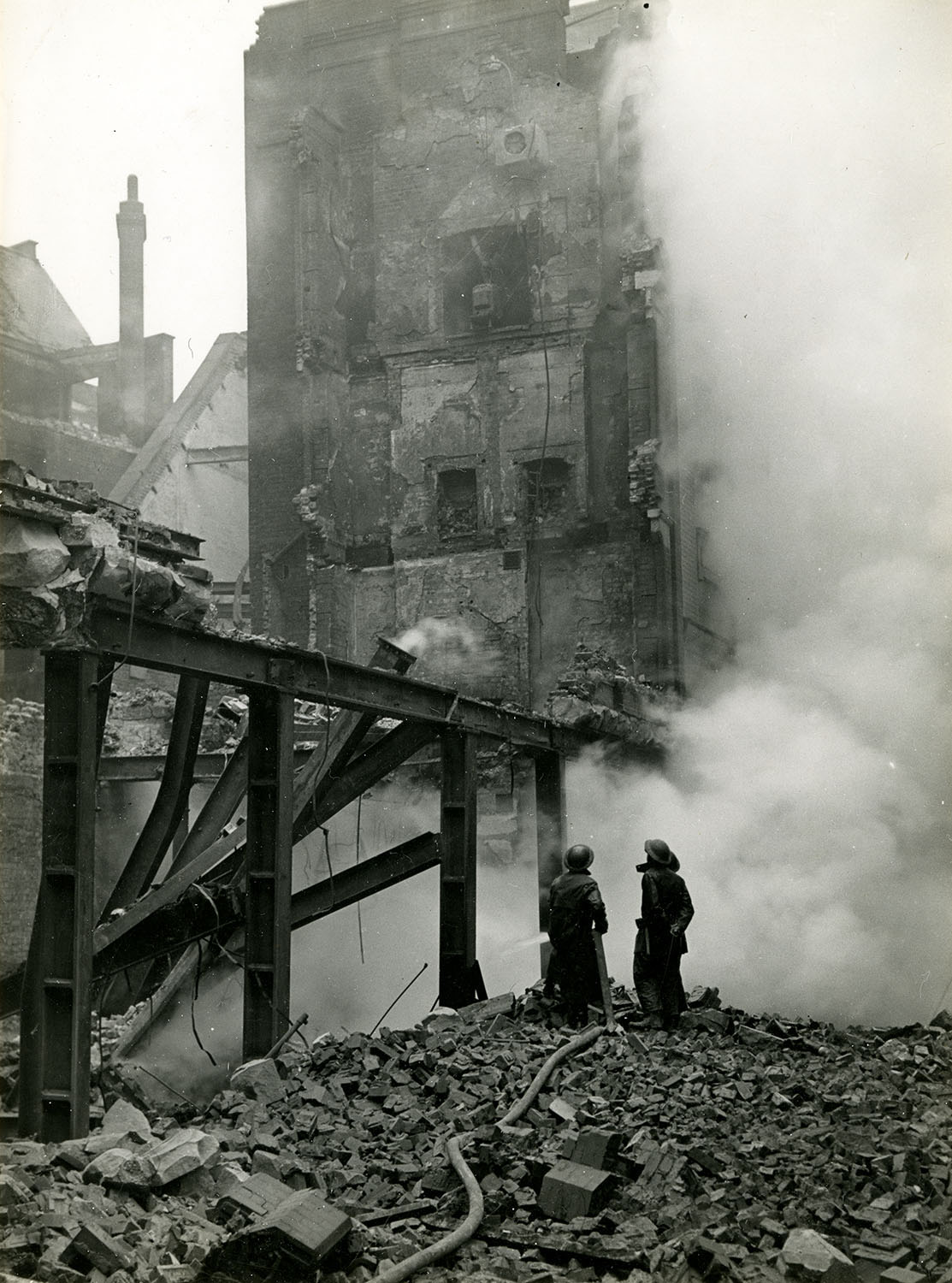Two fire fighters stand on a pile of rubble next to steel girders looking up at a bomb damaged building partially shrouded by smoke.
