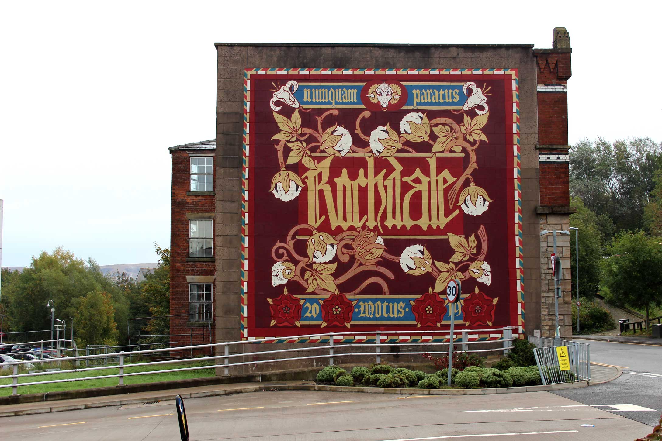 A mural featuring the name of the town, its Latin motto, cotton bolls and red roses, created on the side of a mill building.