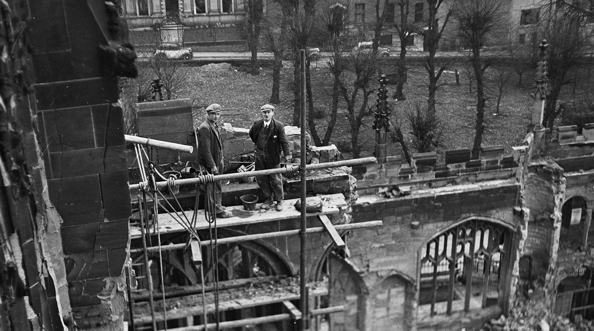 Two men standing high up on scaffolding at the ruins of Coventry Cathedral, look up at the photographer above them in the tower. Black and white photo taken in the 1940s.