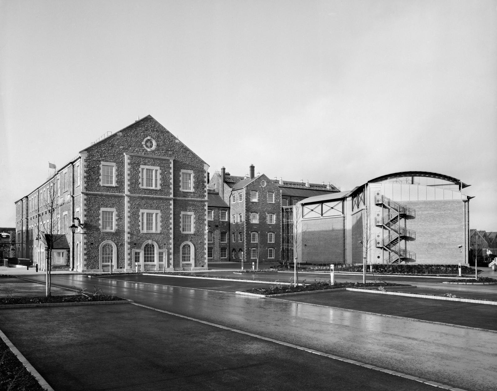Black and white photo of a 4-storey brick office building, with an empty car park in the foreground.