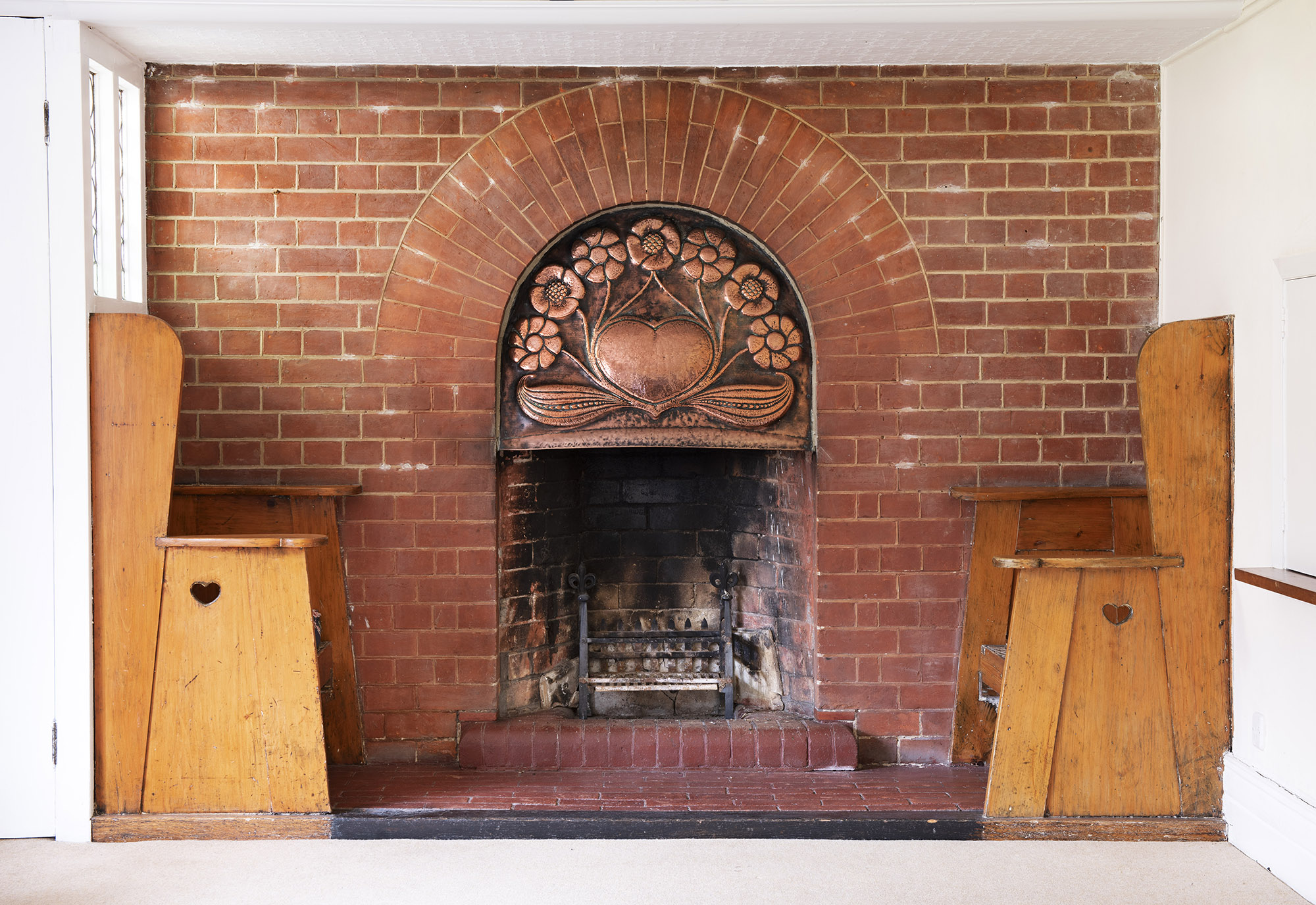 Interior of dining room with the ingle nook on its western wall having fixed seats at either side with cut-out heart shapes. The hearth has a semi-circular opening and a beaten copper fire hood. The copper fire hood has a central heart surrounded by flowers and leaves, emblematic of the Immaculate Heart of Mary.