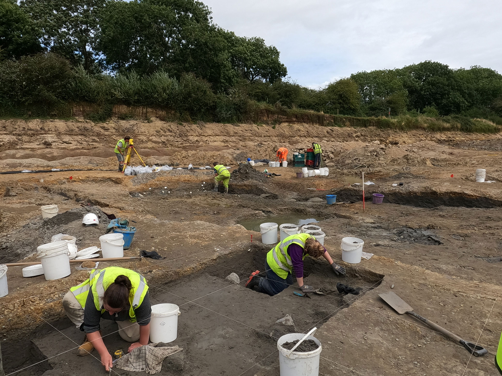 A group of archaeologists excavating: in the foreground two archaeologists are working within a 1 metre grid marked by strings.