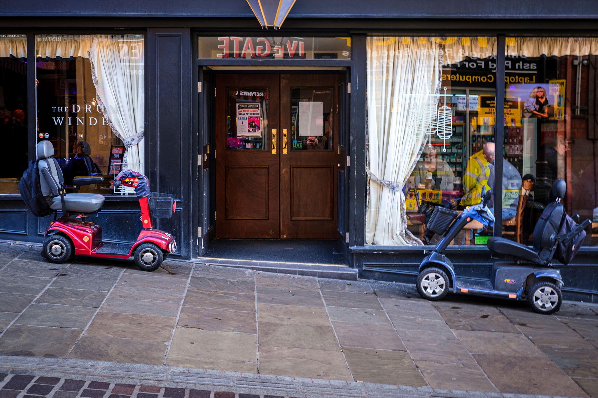 A red mobility scooter and a blue mobility scooter parked on a sloped pavement outside a shop.