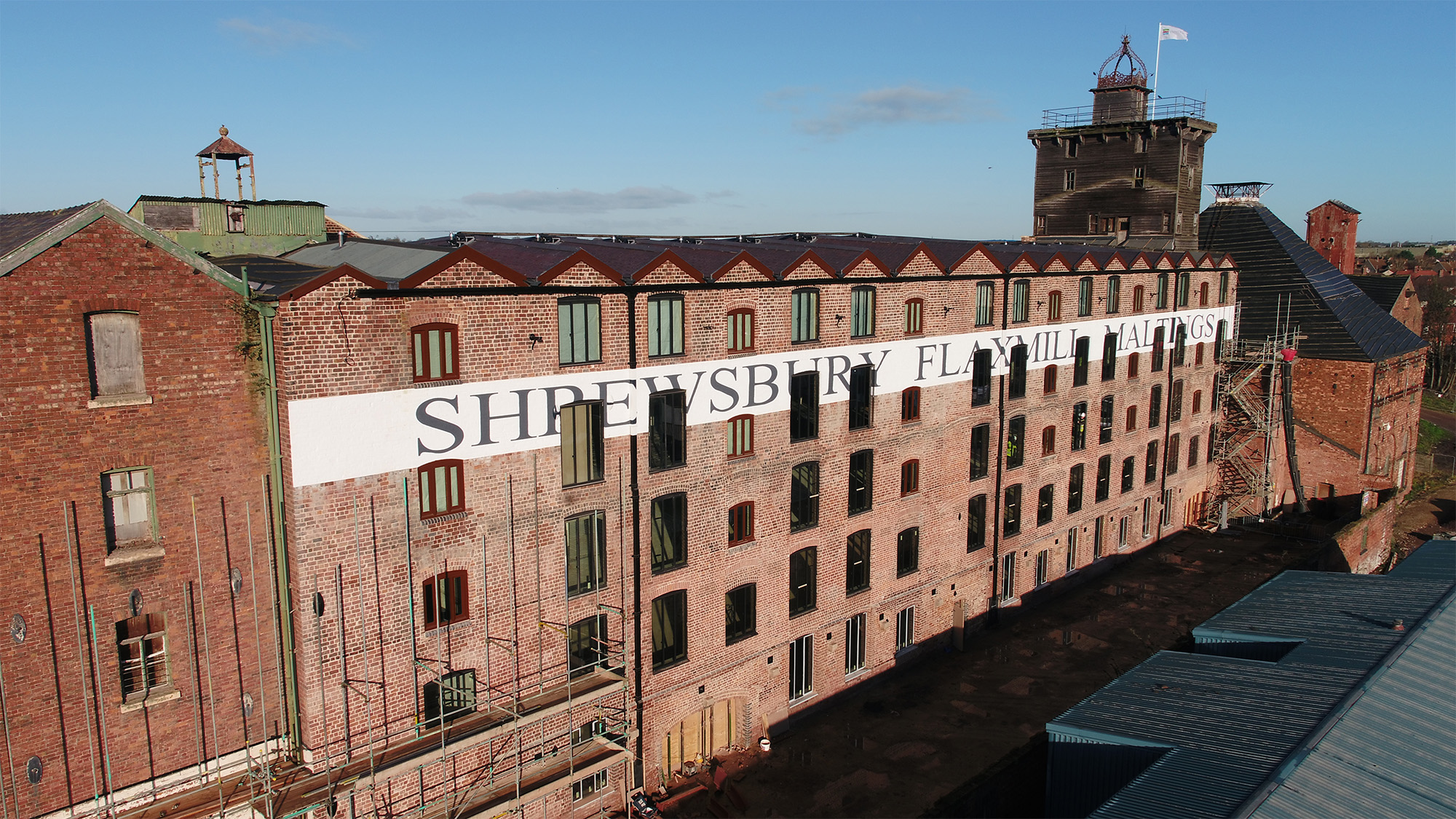 Elevated photo of the exterior of a red brick 5-storey mill building with a sign reading 'Shrewsbury Flaxmill Maltings'.