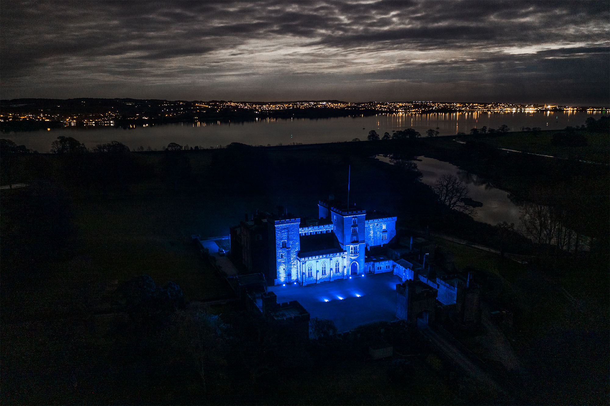 Night-time image showing Powderham Castle lit up blue for key workers against a cloudy sky and the lights of a town reflected in the river.