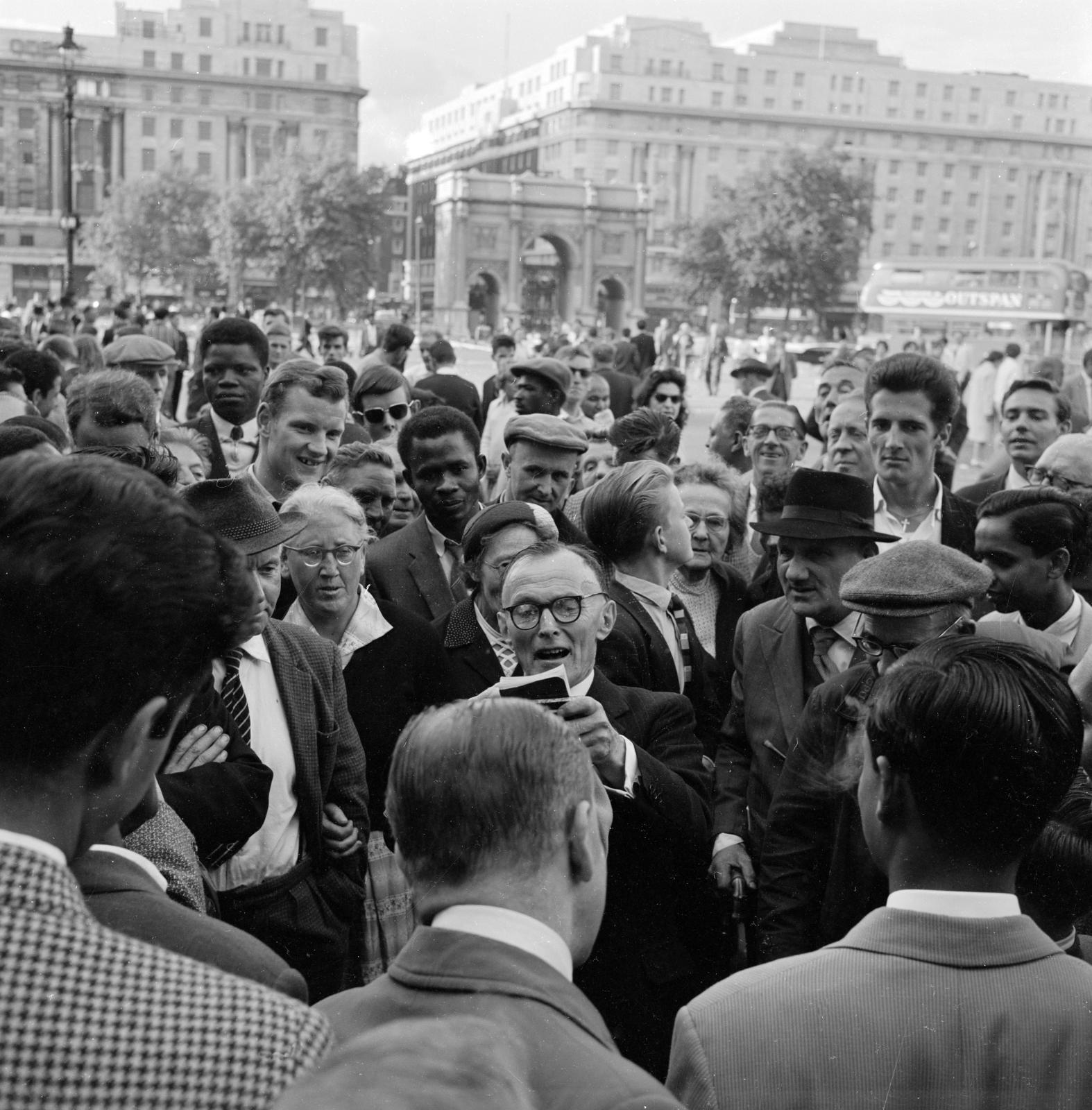 Group of members of the public cluster around a man who is speaking while referring to a notebook.
