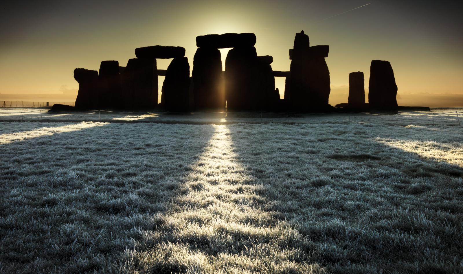 Dawn at Stonehenge with stones silhouetted as the sun's rays create long shadows and shafts of light on the frosty grass in the foreground.