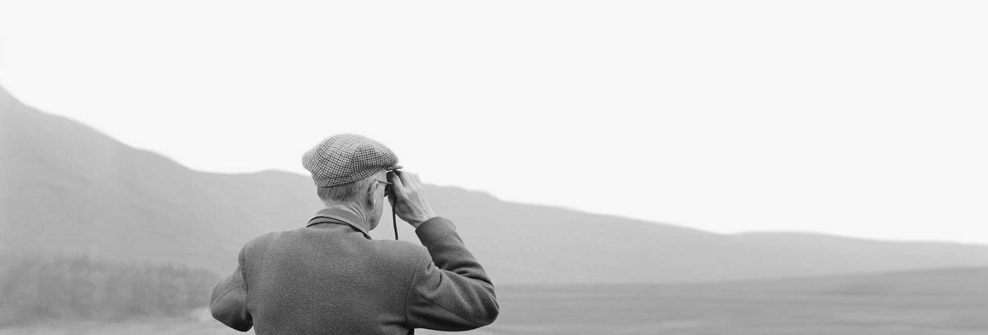 A man standing in front of a dry stone wall using binoculars to look at the fells in the distance. Photographed at an unidentified location near Rydal Water or Grasmere, in the South Lakeland district of Cumbria
