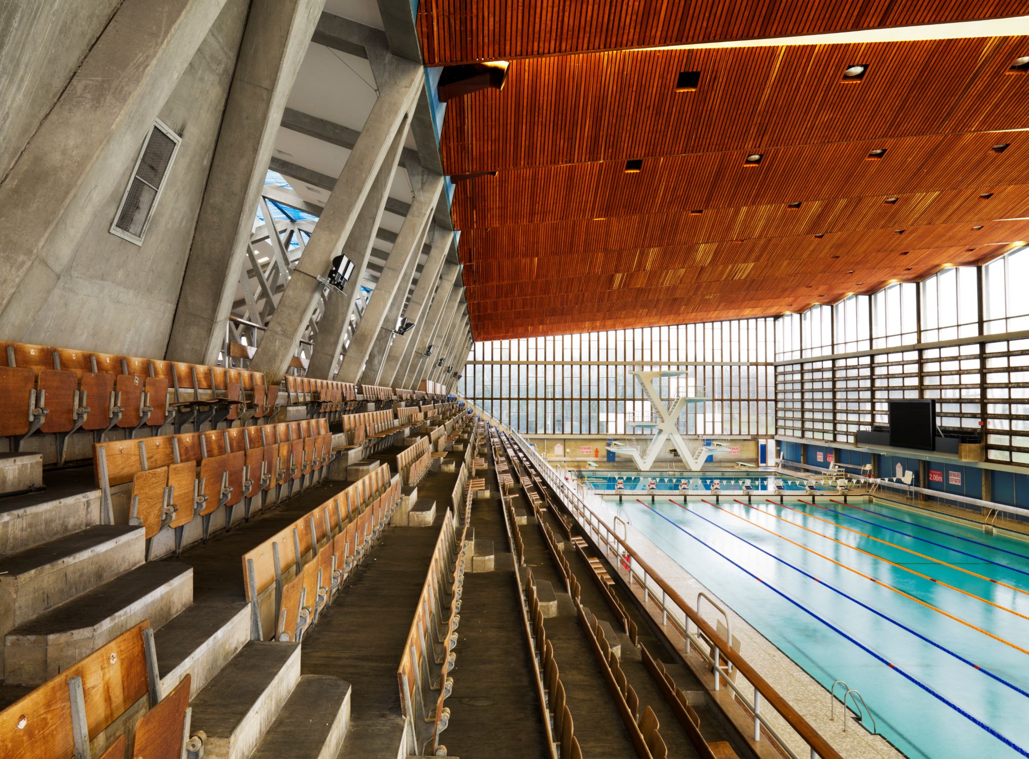 A photograph of an indoor swimming pool with a diving platform and several lanes, flanked by rows of wooden spectator seats. The ceiling features warm-toned wooden panels, and large windows allow natural light to illuminate the space.