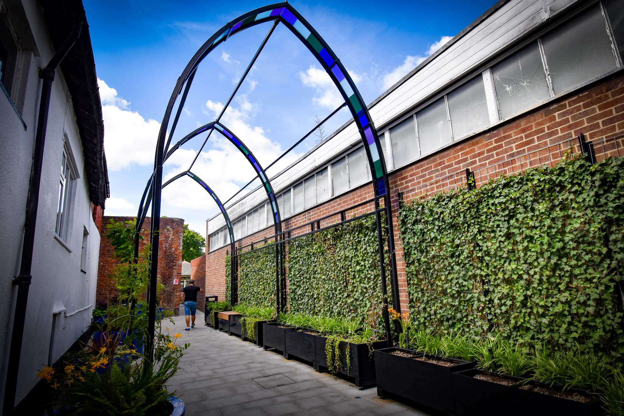 An alleyway lined with planters and an arched metal and coloured glass pergola.