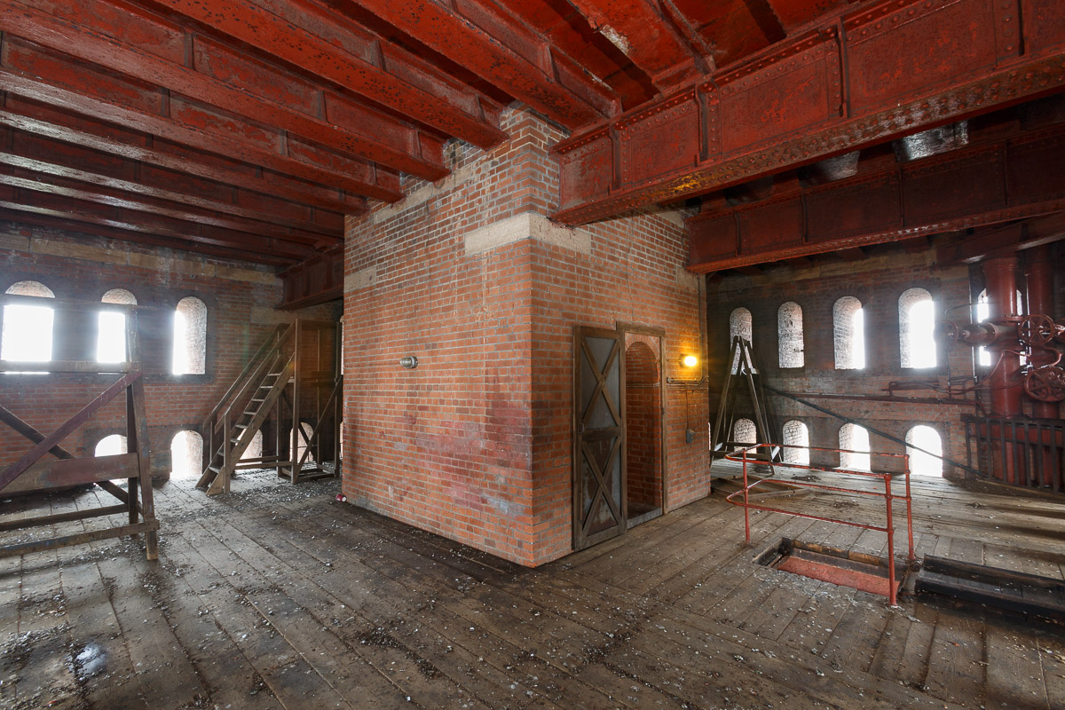 Large room with wooden floor, metal beams in the ceiling, two levels of arched windows and a large brick casing with door leading to stairwell.