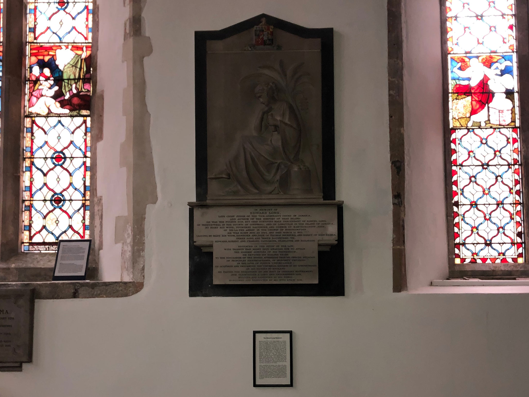 A Neo-classical memorial with a relief sculpture and plaque situated on the wall of a church between two stained glass windows, below the memorial is a simply framed modern text document.