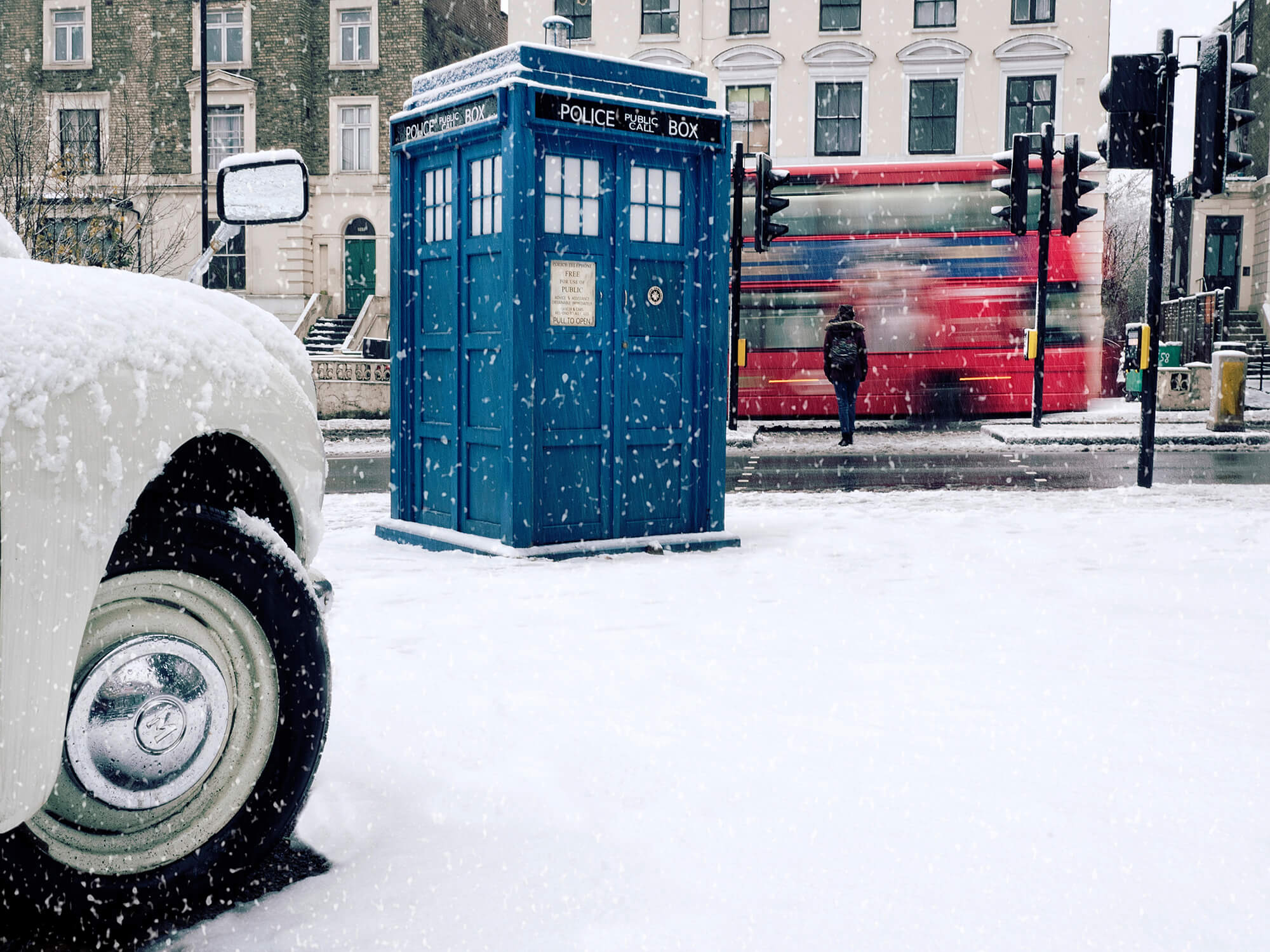 Tardis on snow covered pavement next to pelican crossing with pedestrian walking away over the crossing, pausing as a red double-decker bus passes. On left in foreground is the front right wheel an arch of a white car.