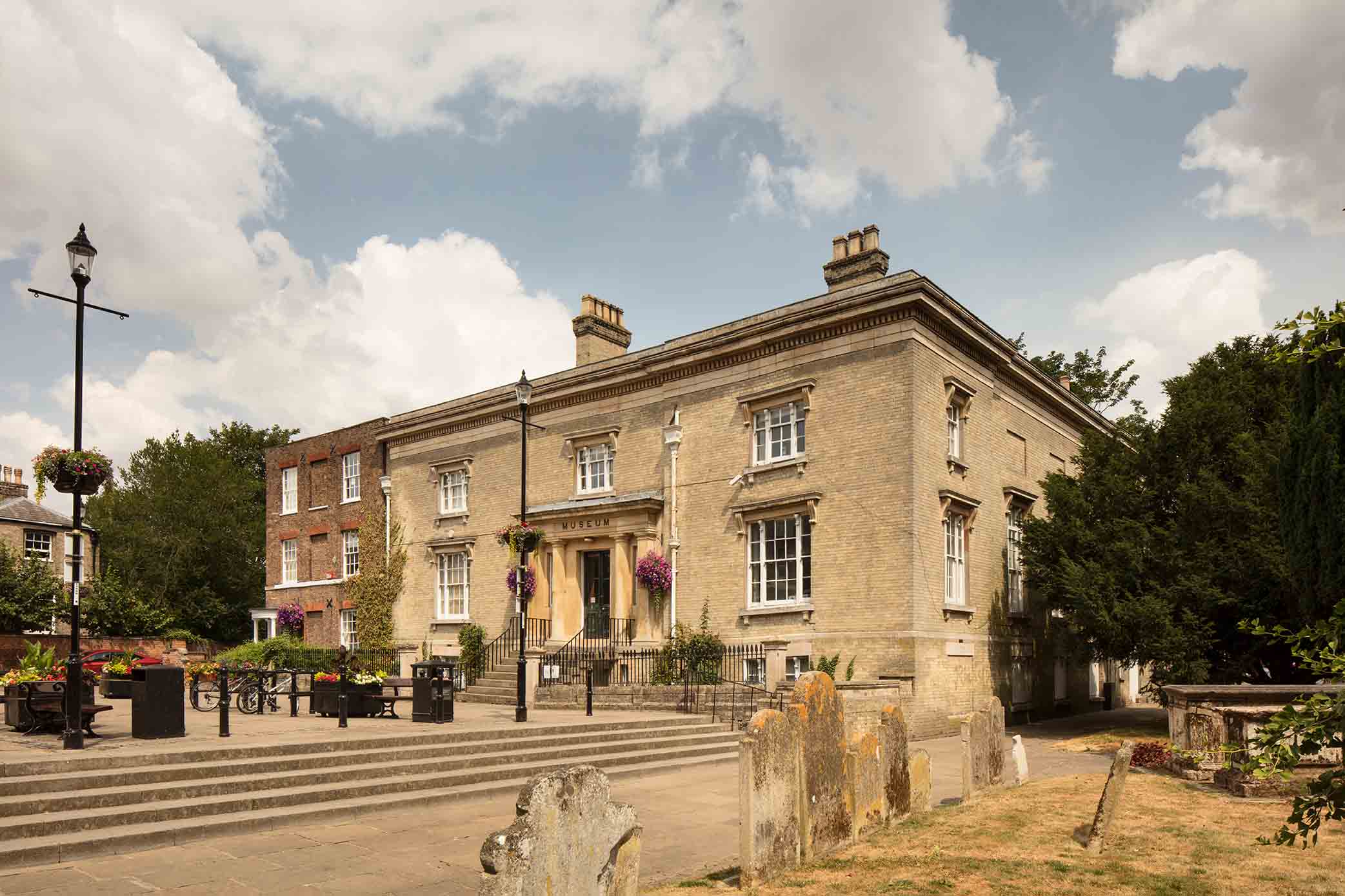 An old graveyard to the right and then steps leading up to antique wrought iron lamp posts and a sandy coloured brick building with front porch and pillars.