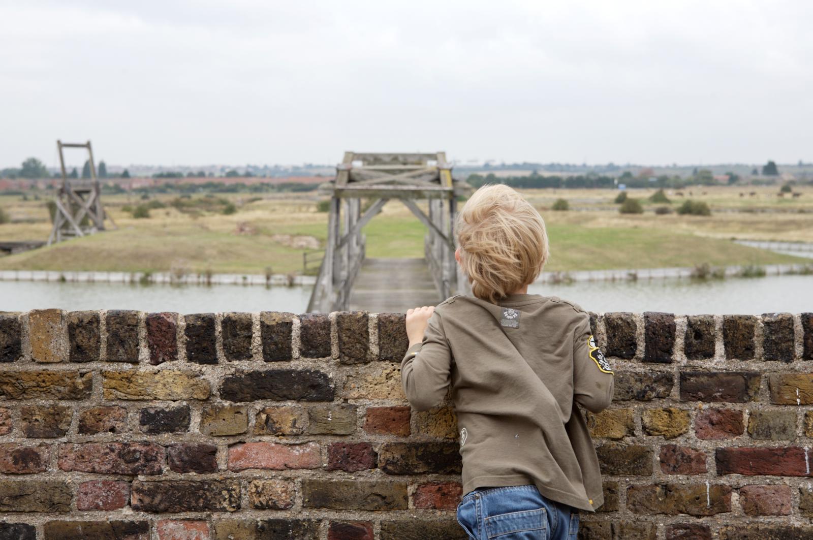 Boy looking over fort wall to wooden bridge in distance.