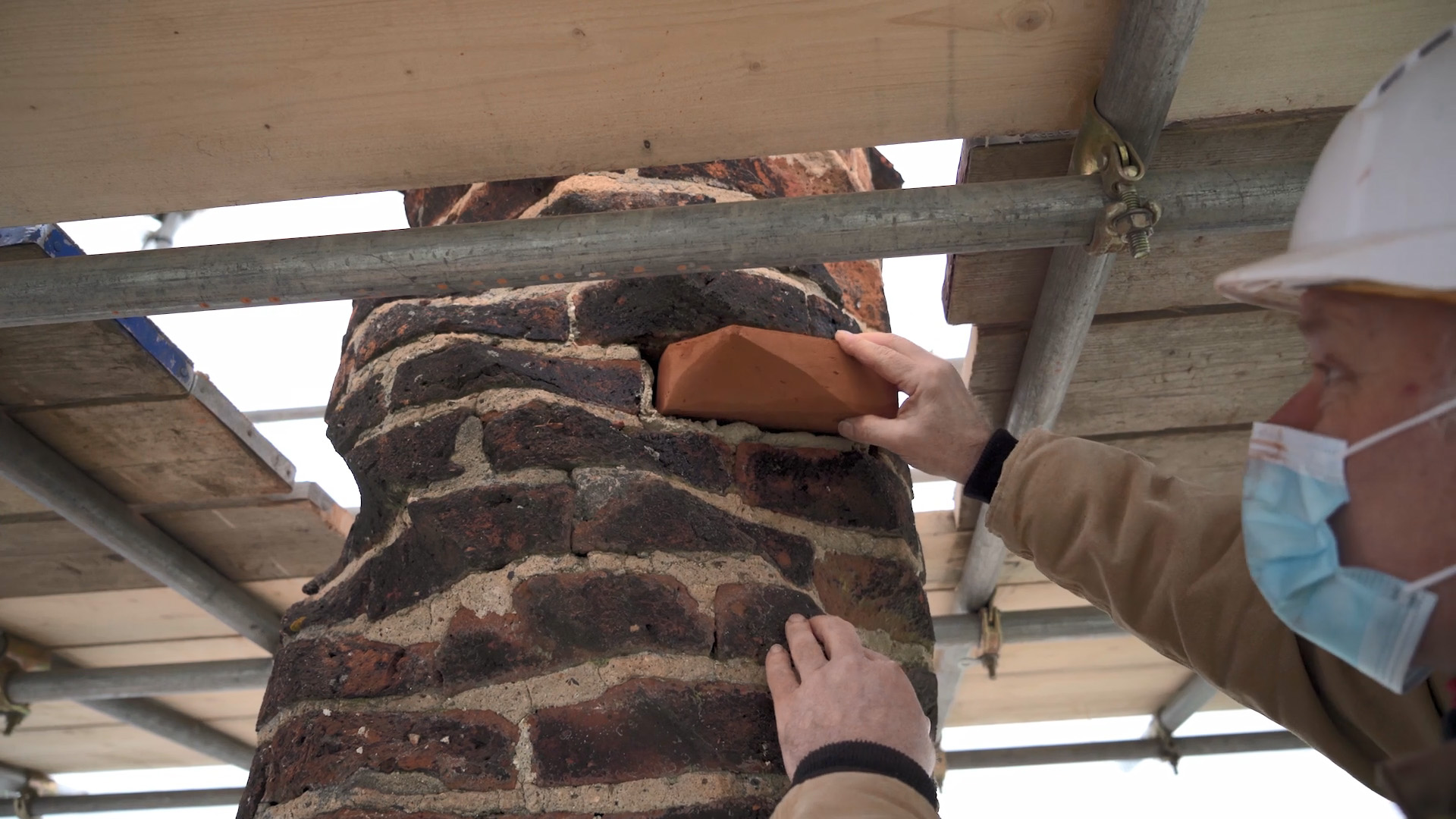 Heritage craftsman replaces a hand-carved brick in a Tudor brick chimney, that is sculpted as if it is twisting around. The craftsman is wearing a facemask and the chimney is surrounded by scaffolding.