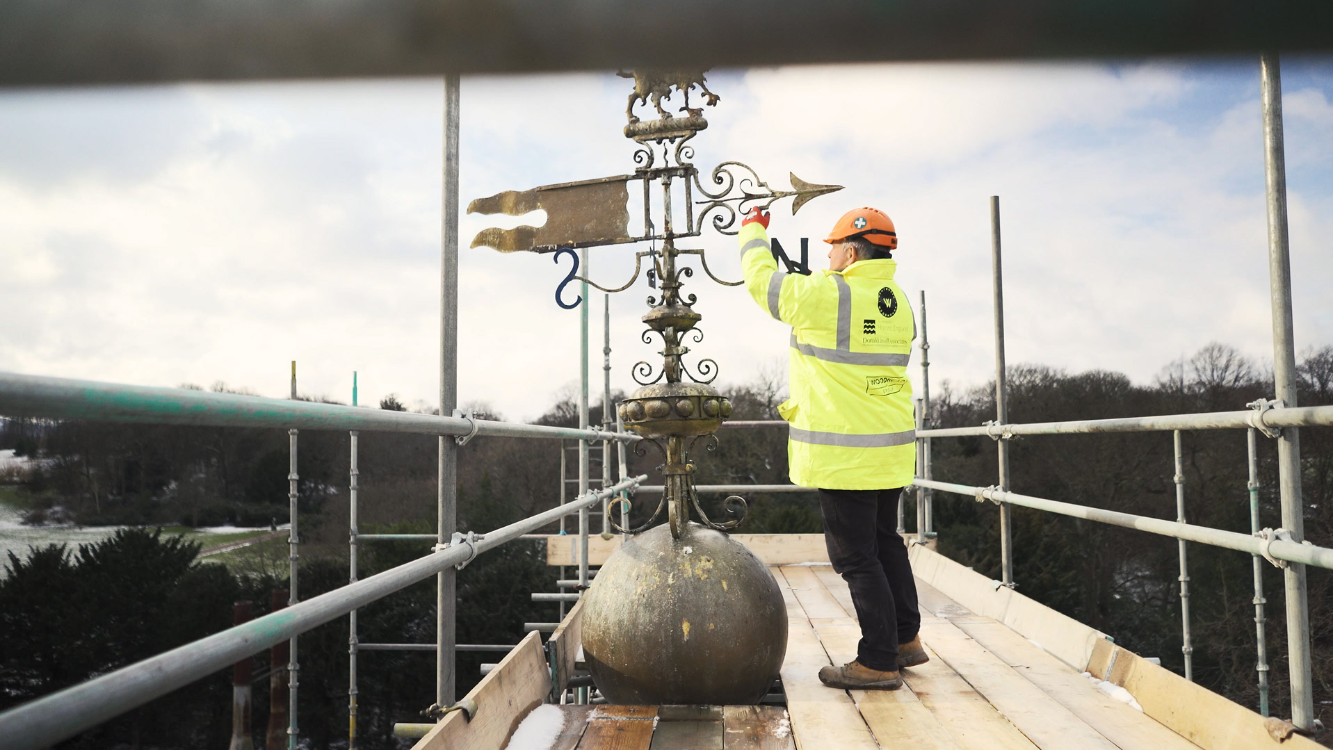 Heritage craftsman in a hard hat and high visibility jacket stands on scaffolding on the Wentworth Woodhouse, and reaches up to the weathervane. The sky behind him is cloudy.