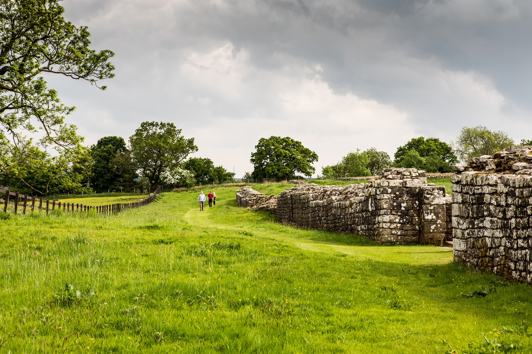People walking on grass alongside a long stone wall
