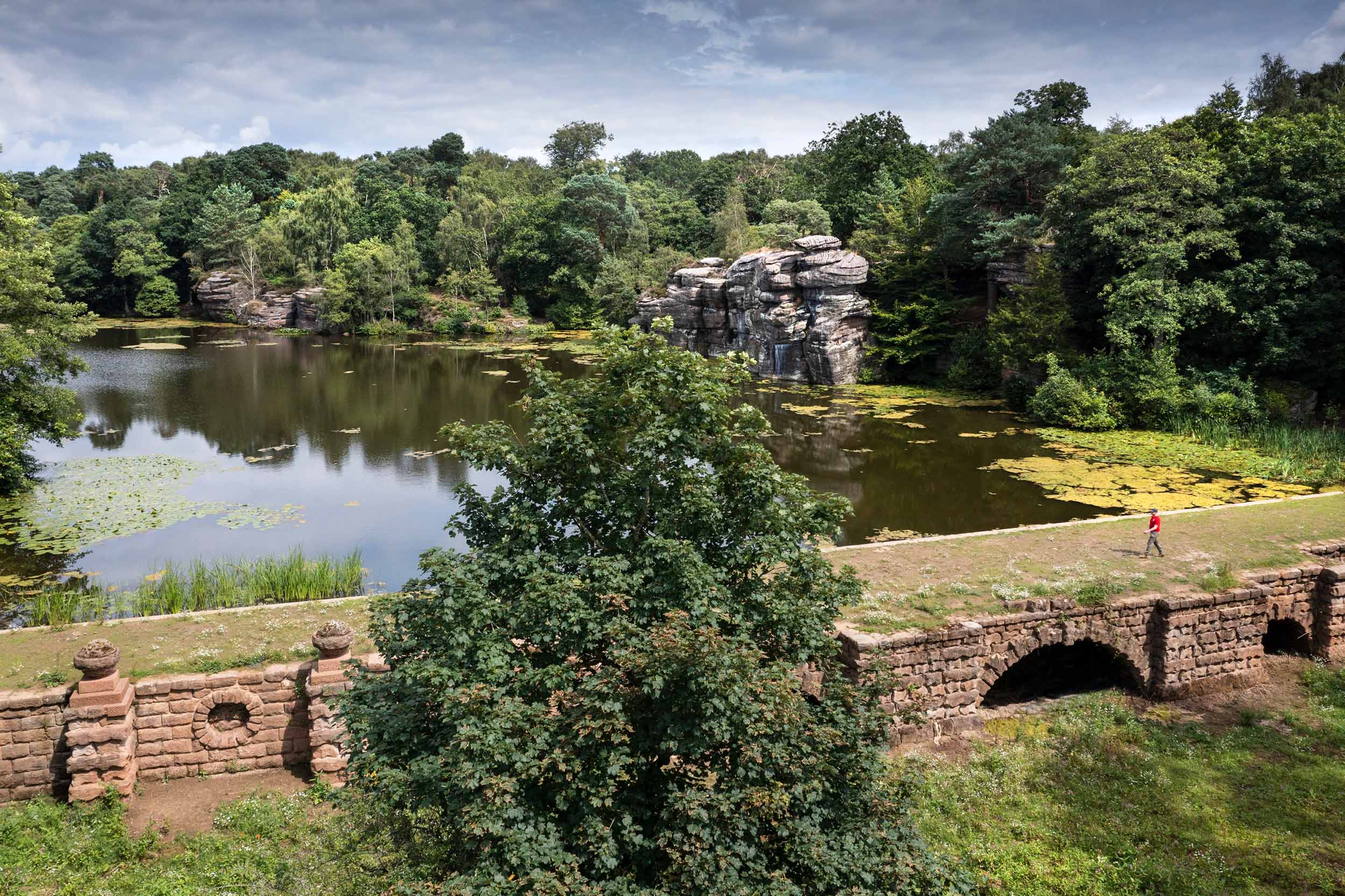 Landscape gardens, lake, rocks and dam wall at Plumpton Rocks. A man walks along the dam wall on the right of the image.
