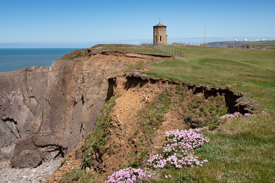 A historic tower stands perilously close to the edge of an eroded cliff in a coastal setting.