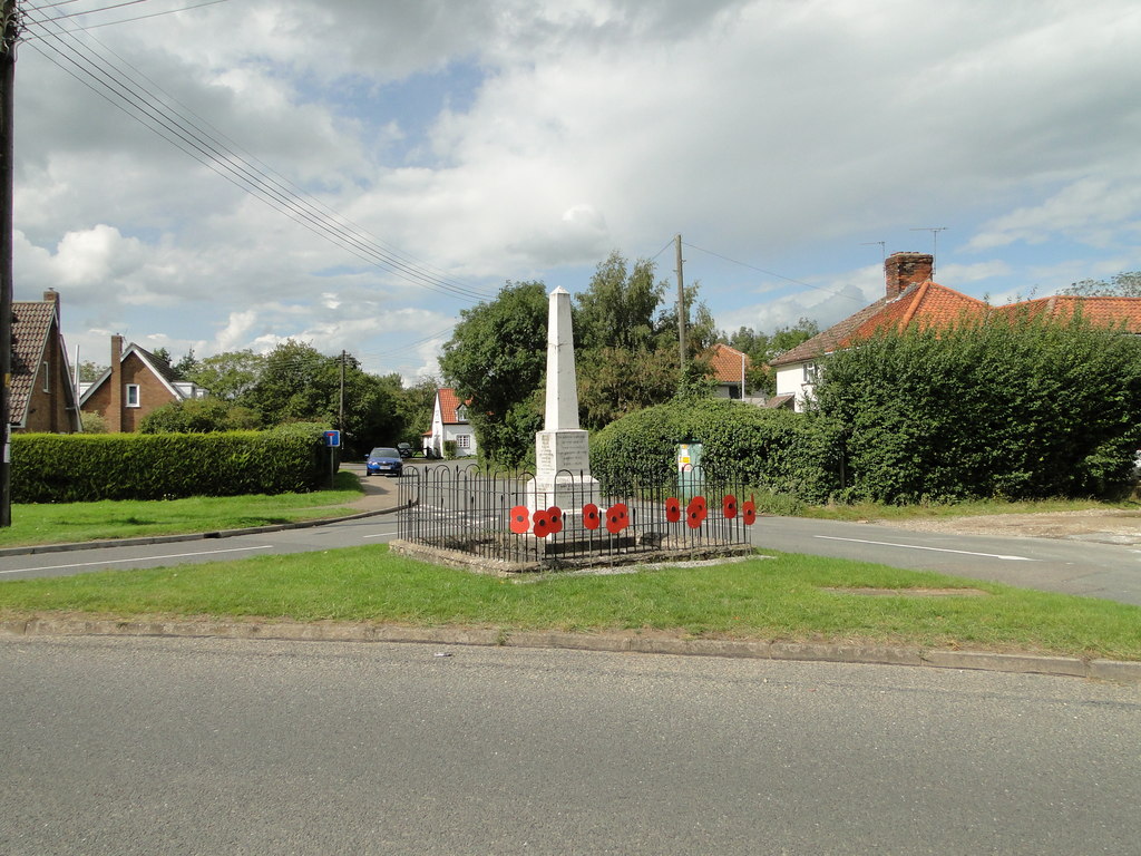 A wide-angle photo of a war memorial on a triangular grass verge at a road intersection