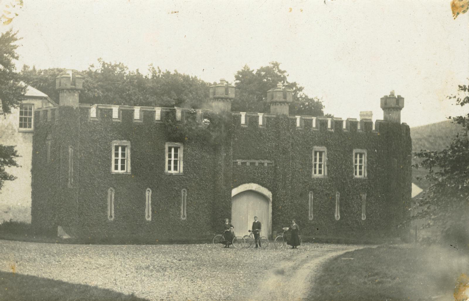 Black and white photo of an unidentified ivy-clad, crenellated 19th-century gatehouse with three cyclists outside main entrance.