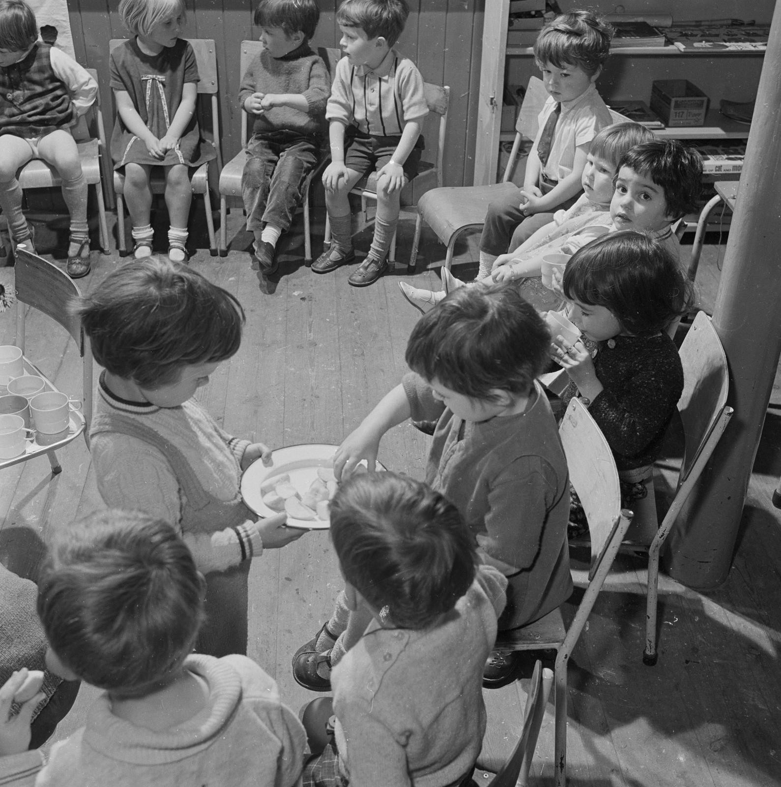 Children sitting in a circle as one standing child offers one of the seated kids a plate of food.