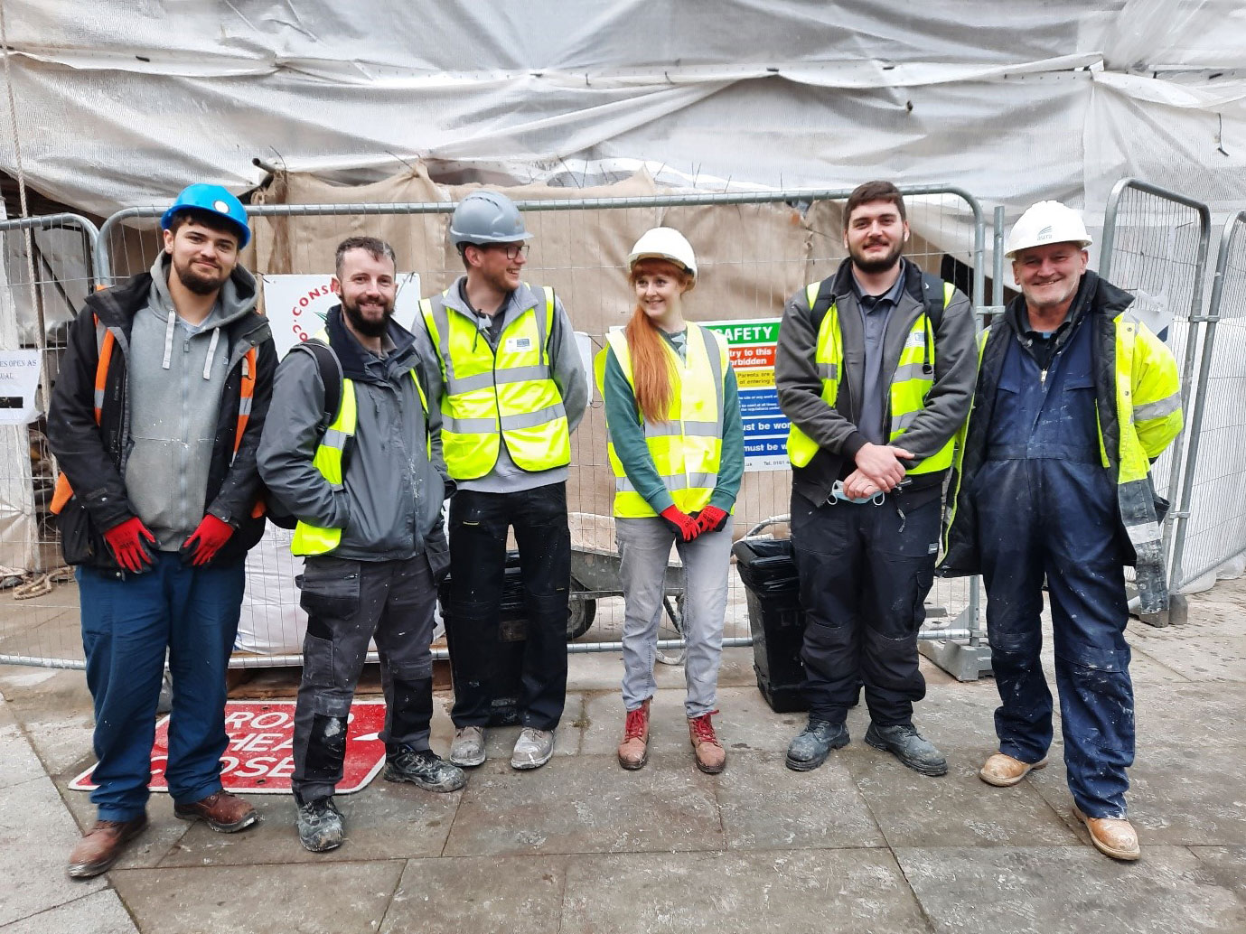 A group of apprentices and other staff in construction PPE pose in front of a building shrouded in plastic sheeting and scaffolding.