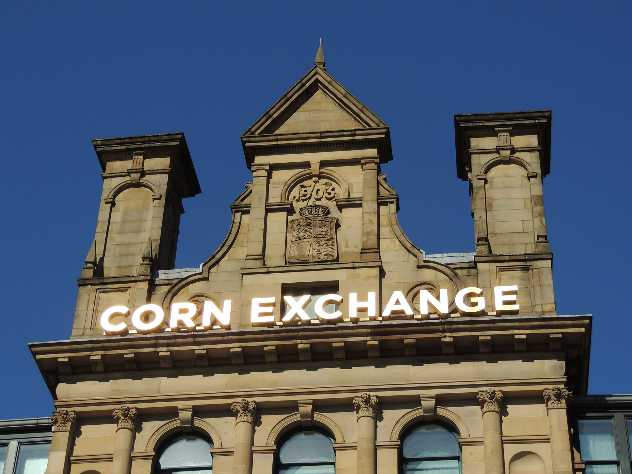 A close-up photo of the top of a yellow-stone building, focusing on columns, illuminated letters spelling 'CORN EXCHANGE', and a coat of arms with '1903' carved above.