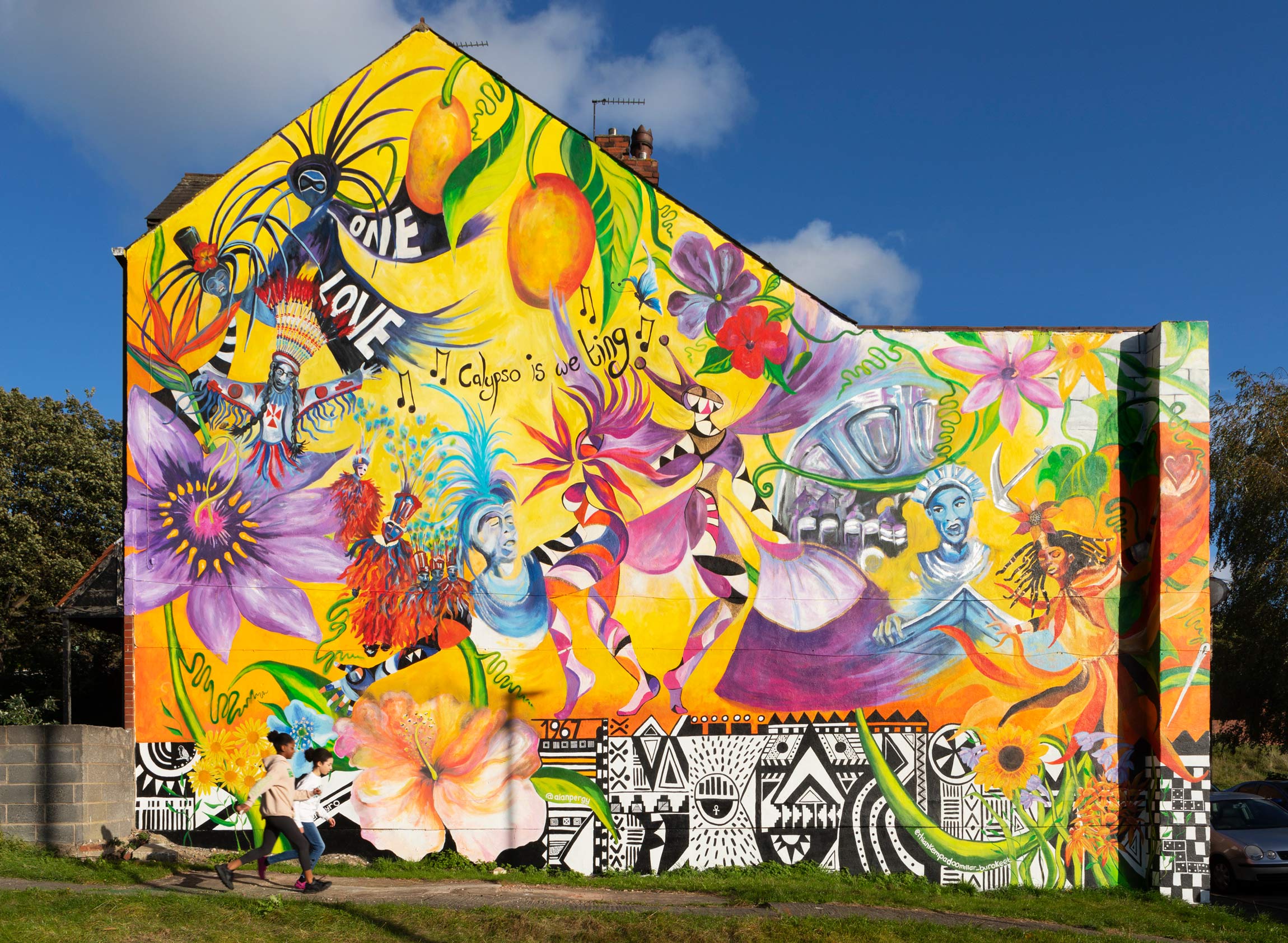 Colour photograph showing part of a mural painted on the whole of the side elevation of a house. The mural features flowers, fruit and depictions of people in costume. Text within the mural includes: 'One Love' and 'Calpso is we ting'. In the foreground is a narrow path between strips of grass. Two children run along the path from the left.