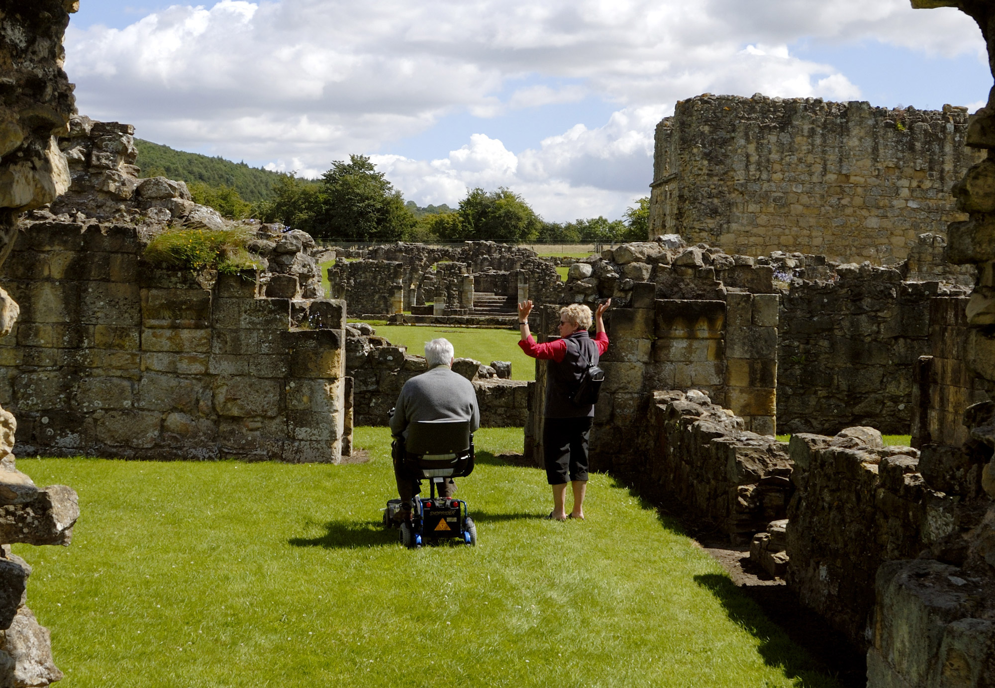 2 people, 1 standing and 1 seated on a mobility scooter, surrounded by ruined remains of an abbey.