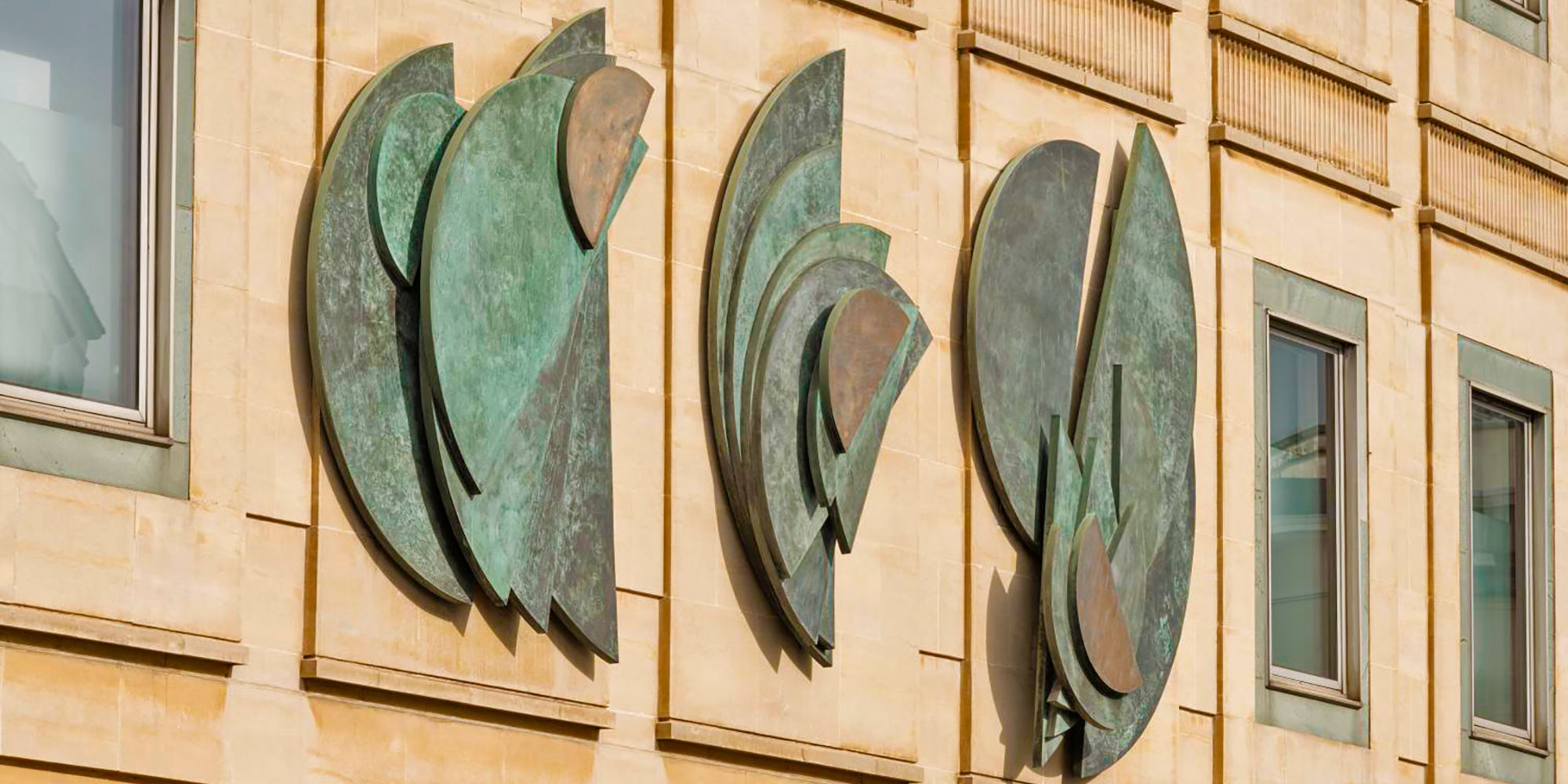 A series of semi-circular bronze sculptural panels adorn a sandstone wall. 