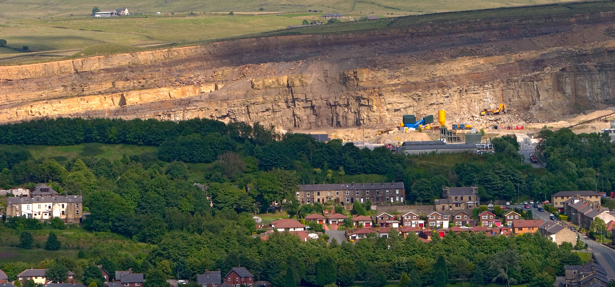A wide view of a broad quarry face, quarry machinery, woodland and rows of houses.  
