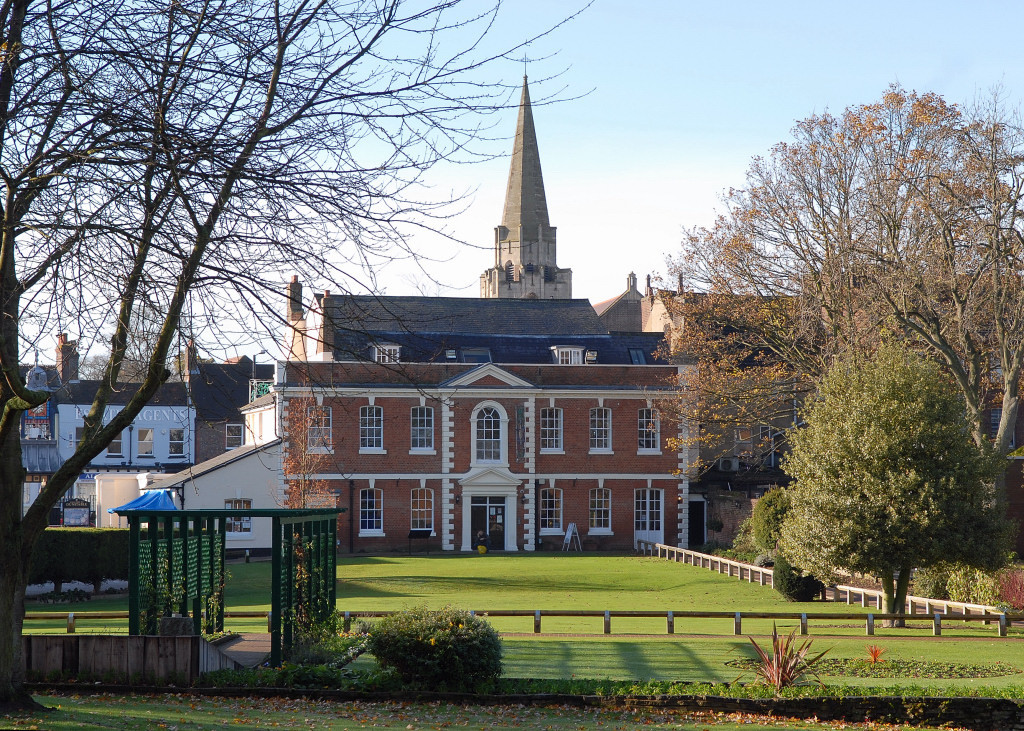The photography shows a green and beyond that the brown brick building of Priory House with white, pillared, arched doorway.