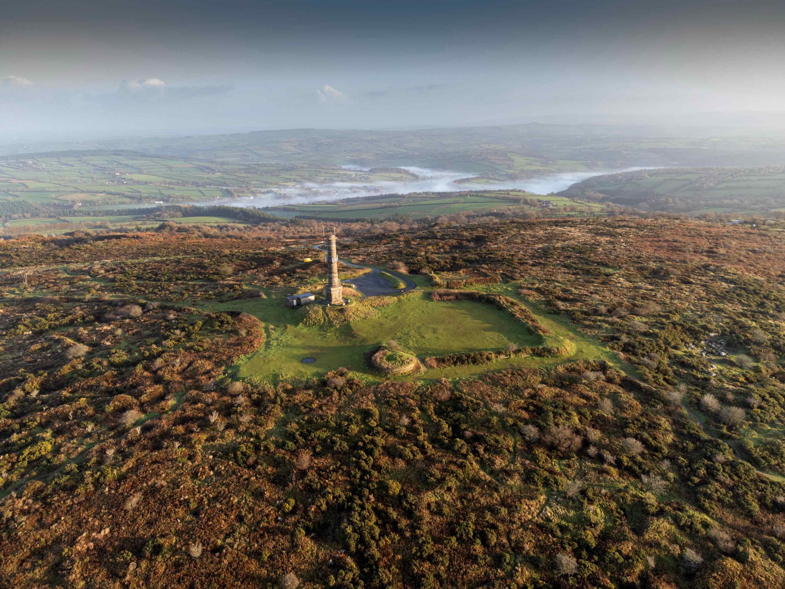 Aerial view from south across the site of Kit Hill, Cornwall. It shows a folly in the centre surrounded by the countryside.