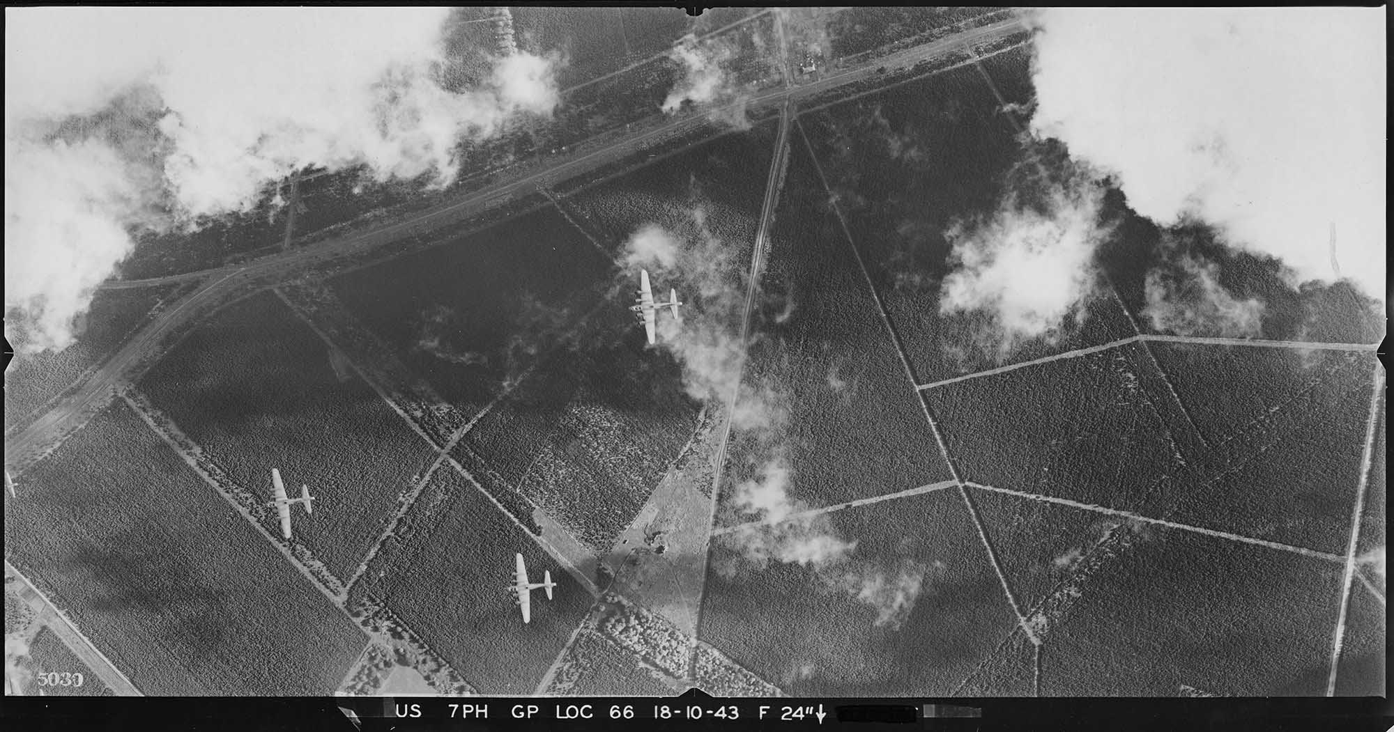 A black and white vertical aerial photograph of three military aircraft flying over a patchwork of plantations. Clouds obscure parts of the photograph and cast shadows on the ground.